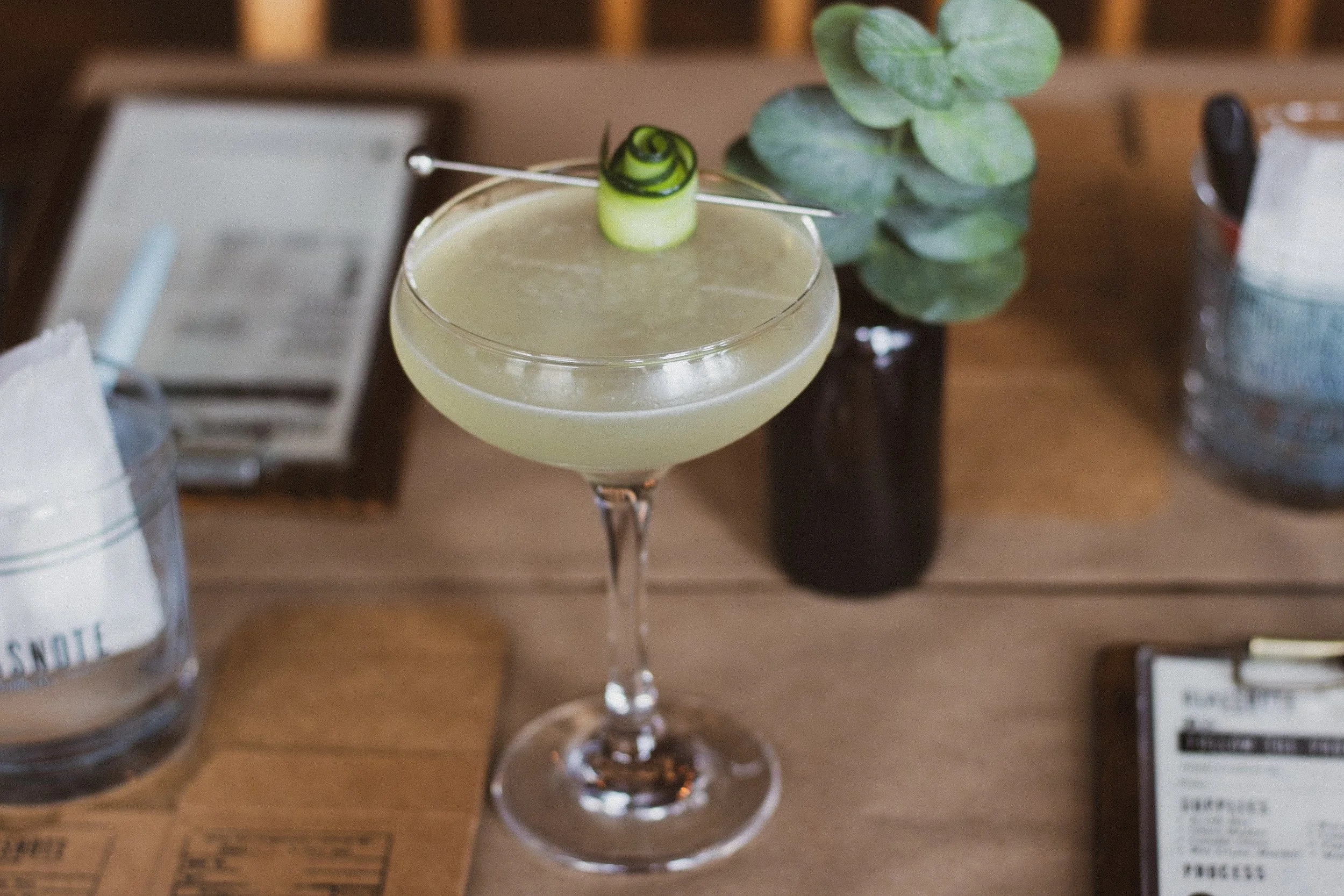 A pale green cocktail in a coupe glass with a spiral cucumber garnish on a metal pick, set on a wooden table with a small potted plant, water glasses, and a menu nearby.