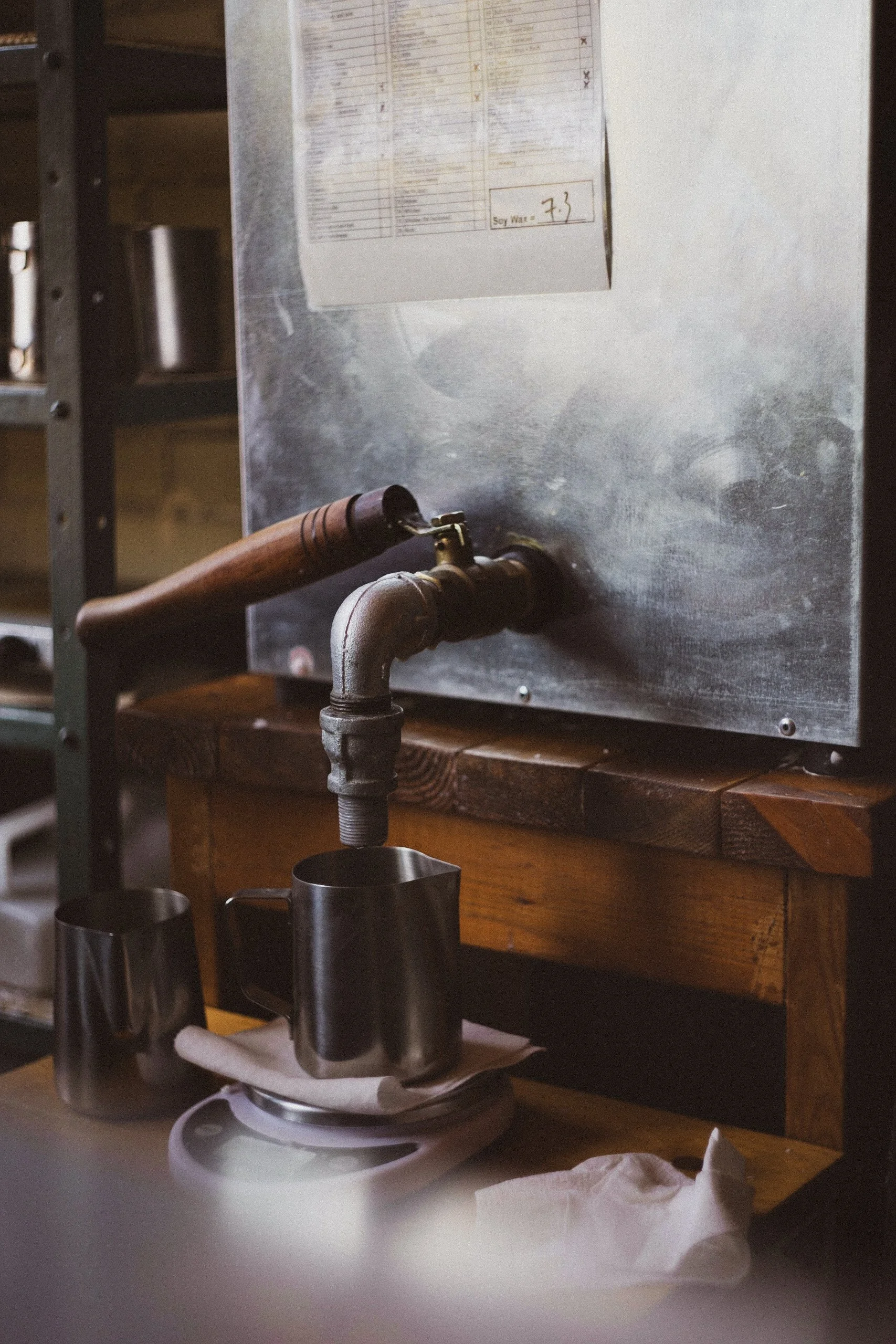 Pouring wax from a metal industrial wax melter into a pouring pitcher. Two additional mugs are nearby, with a paper towel and tissues on the wooden counter.