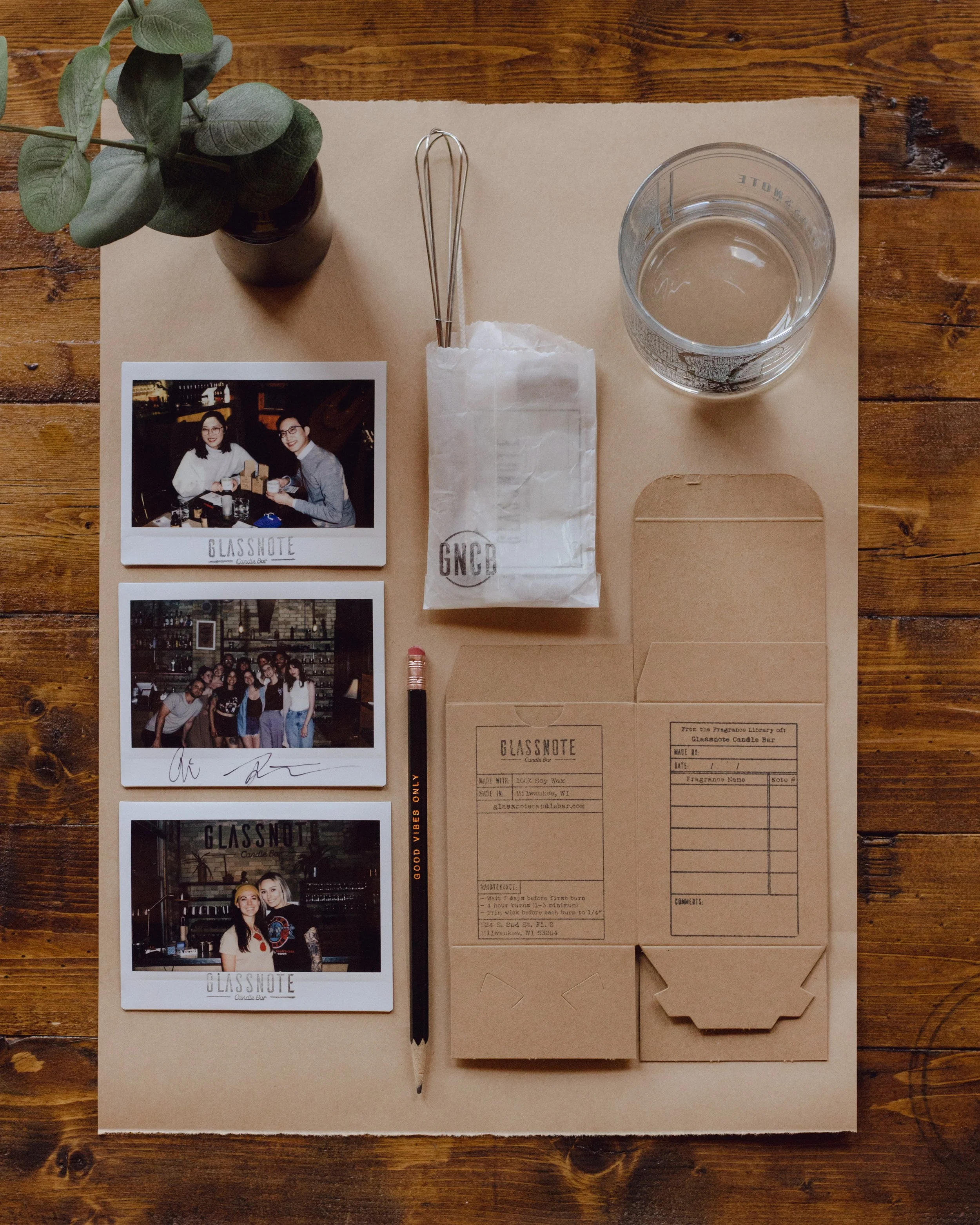 Flat lay of a wooden table with a tan paper mat, holding a small potted plant, three photographs, a black pen, a candle jar, candle making materials, and a cardboard box with printed labels.