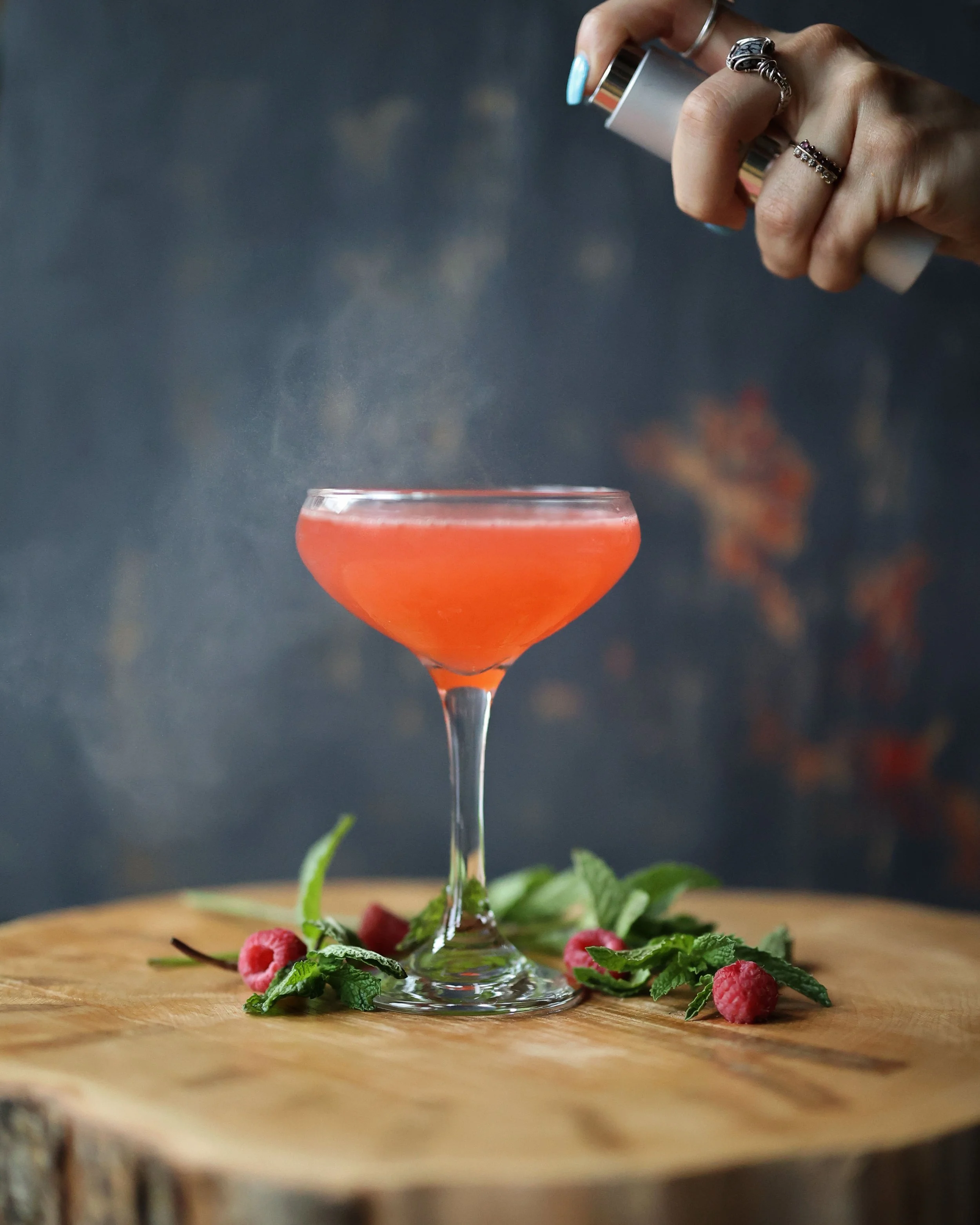 A person spritzes ingredients onto a pink cocktail in a coupe glass, garnished with raspberries and mint, on a wooden surface.