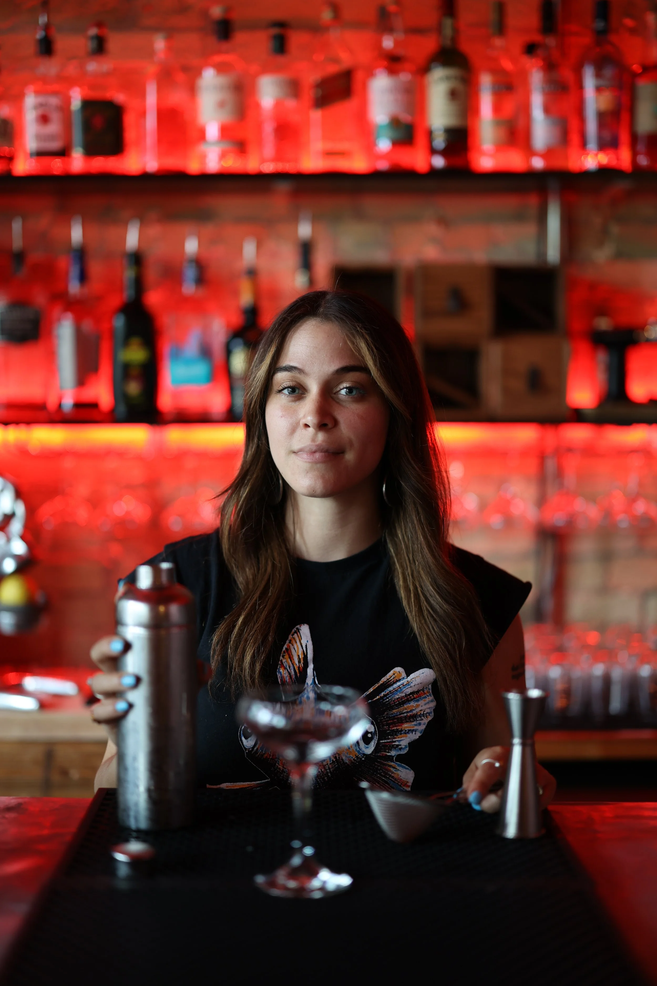 A young woman with long brown hair sitting at a bar counter with cocktail tools, in a bar with red lighting and bottles on shelves behind her.