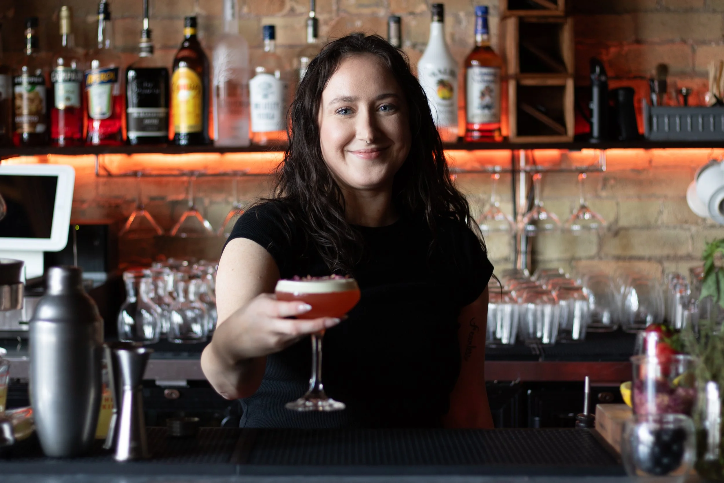 A woman with dark, curly hair smiles as she holds a cocktail in a martini glass at a bar with liquor bottles and glassware in the background.