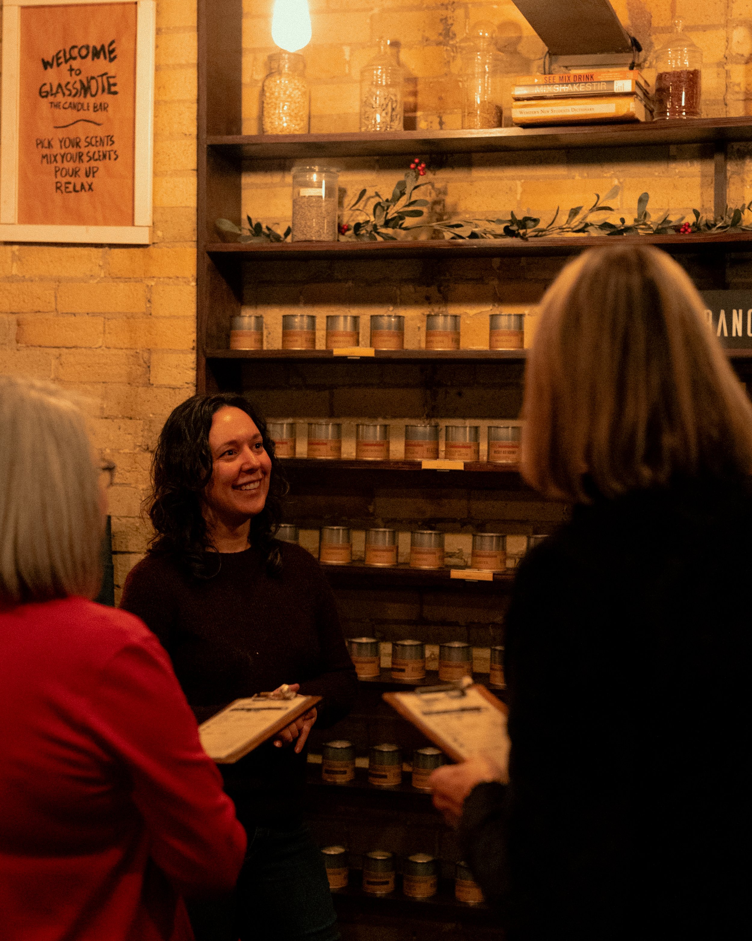 A woman behind a counter smiling while three women with clipboards talk to her inside a candle shop at the fragrance wall with warm lighting, wooden shelves, candles, jars, and books.