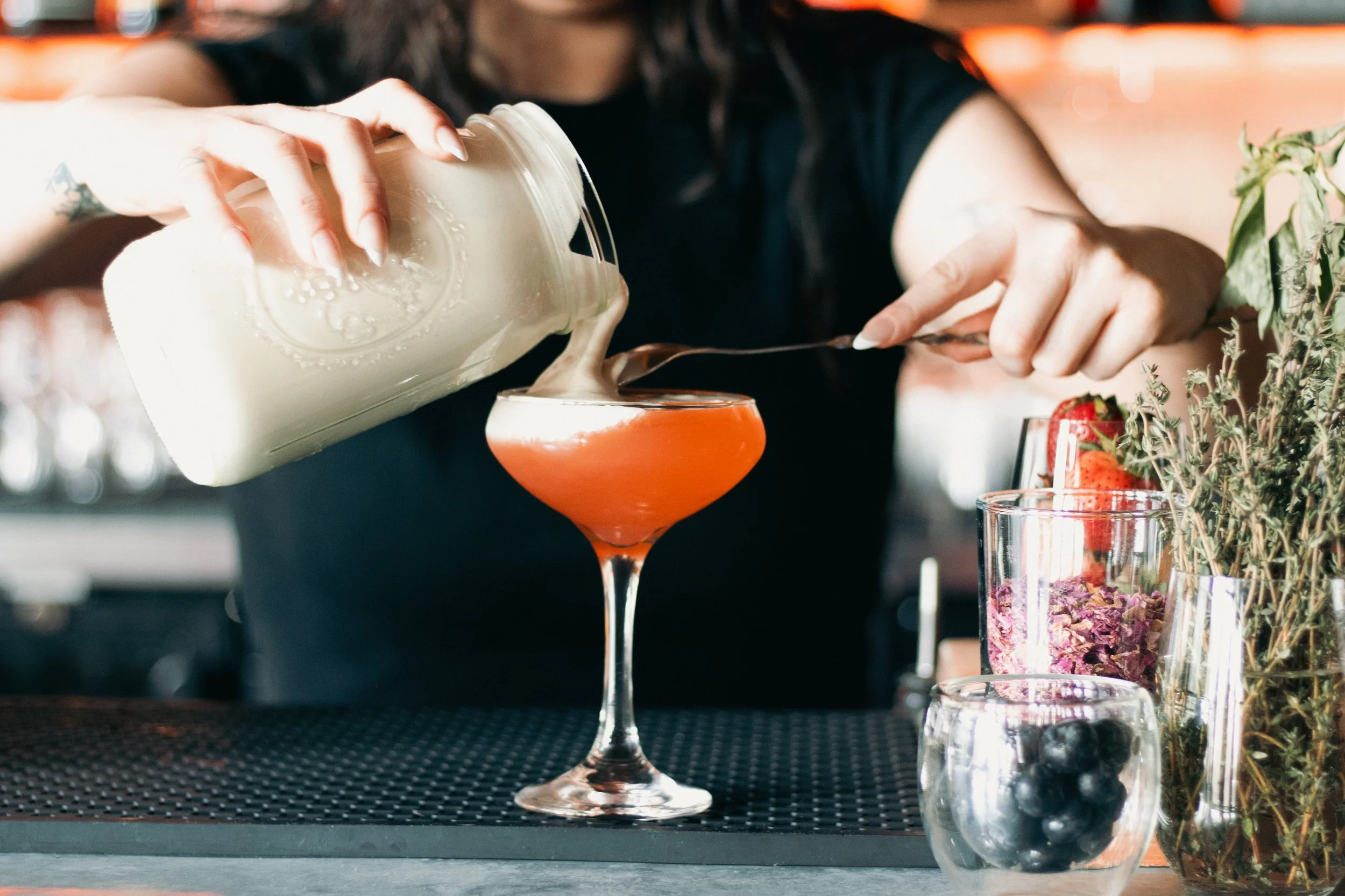 A person in black pouring a creamy liquid from a jar into a cocktail glass with an orange drink, at the zinc bar, with garnishes and plants nearby.