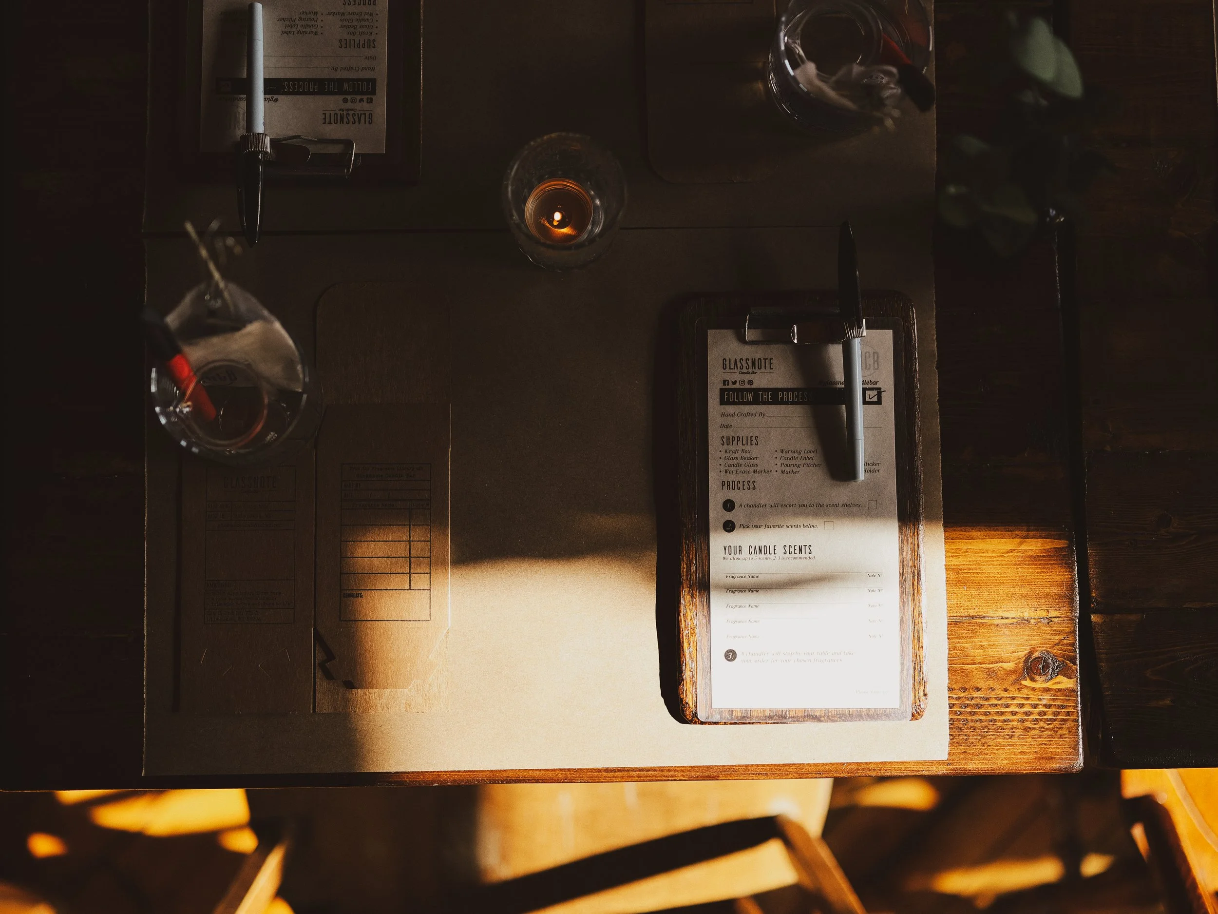 Table setting with a candle, a drink, a glass of water, and a fragrance selection sheet in the warmly lit candle bar. 