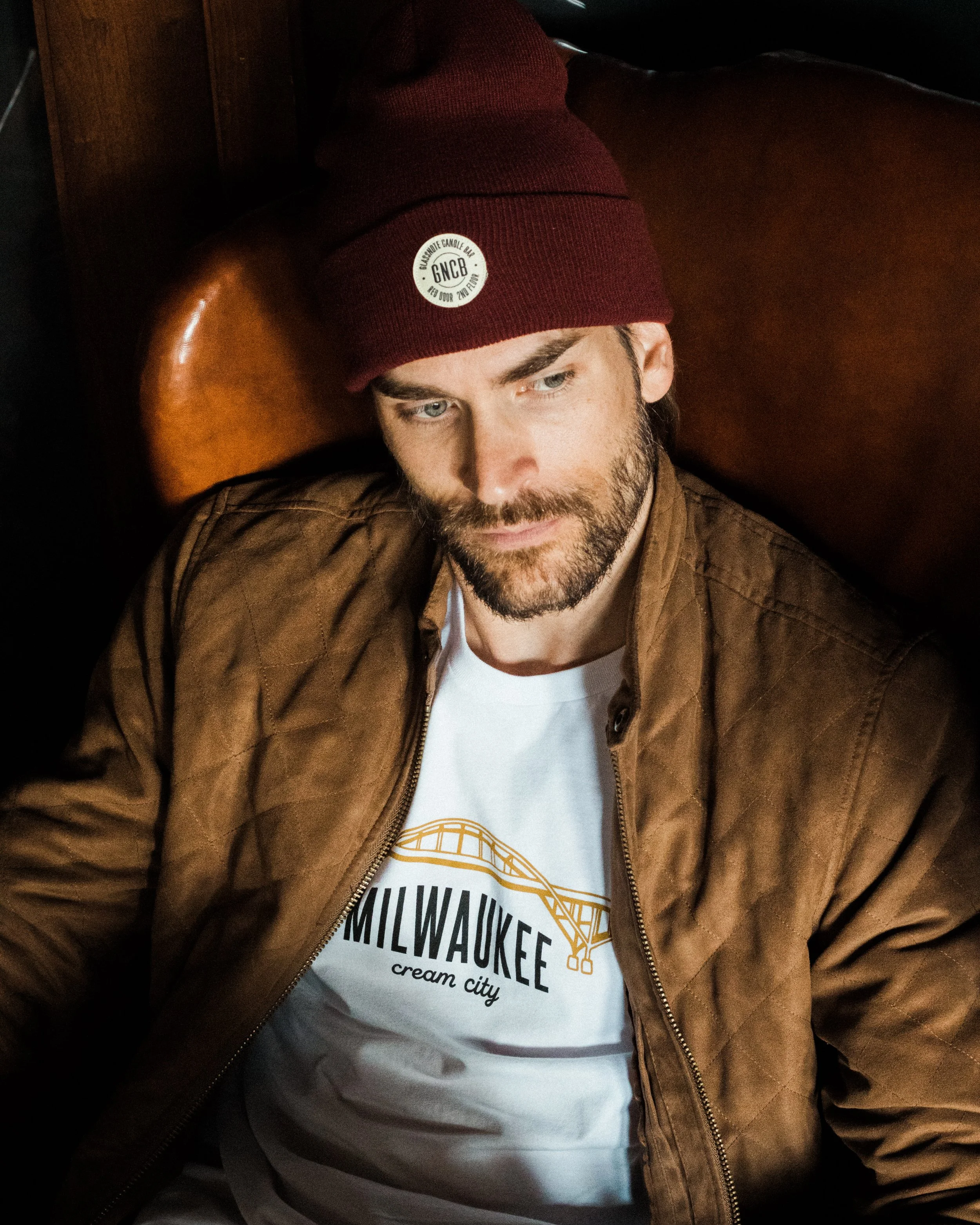 A man with a beard wearing a red Glassnote beanie and a brown jacket, sitting against a wooden headboard, with a T-shirt that has 'Milwaukee cream city' written on it and an illustration of a bridge.