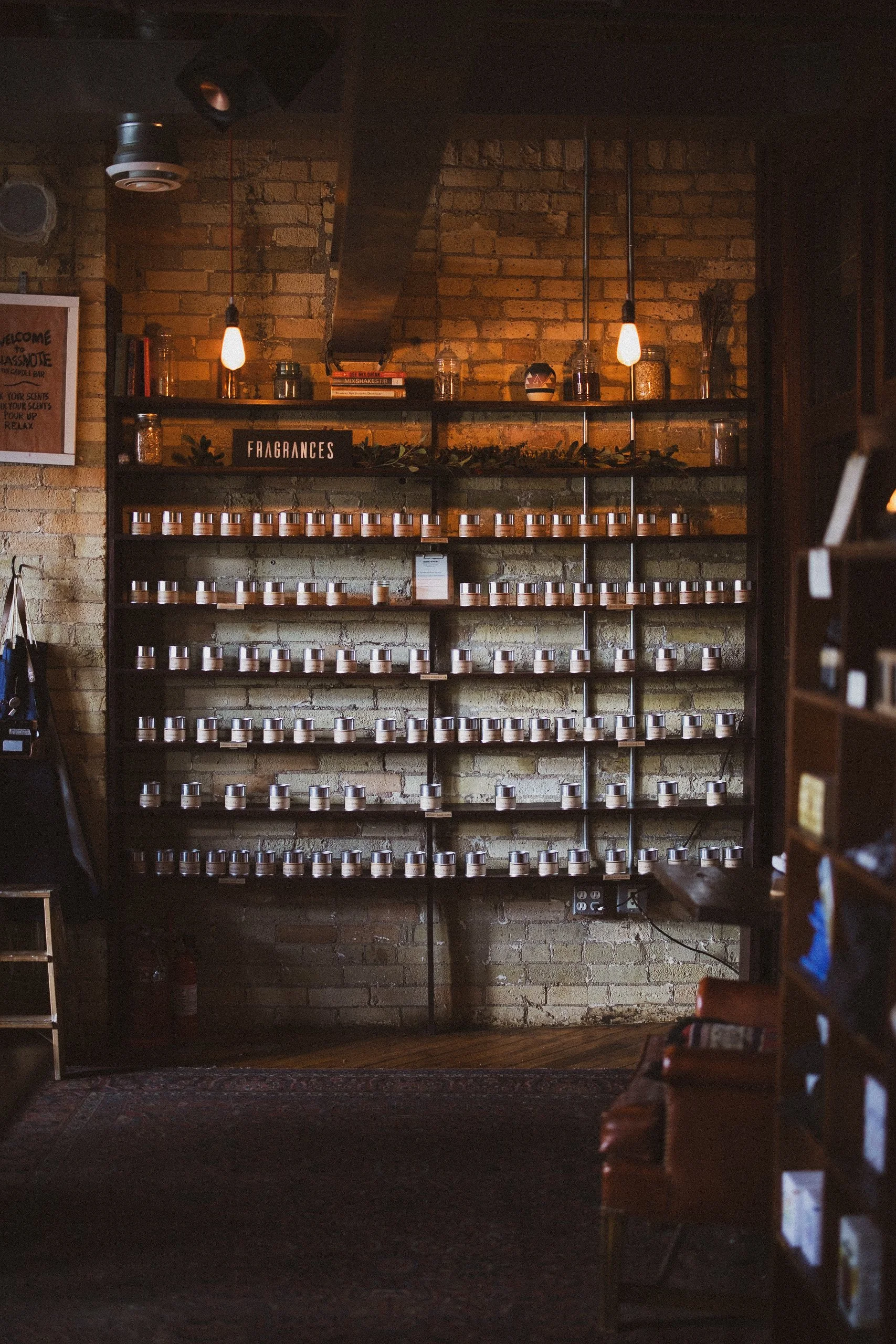 Wall shelf with multiple rows of small jars, labeled "FRAGRANCES," in a cozy candle and cocktail bar with brick walls and warm lighting.