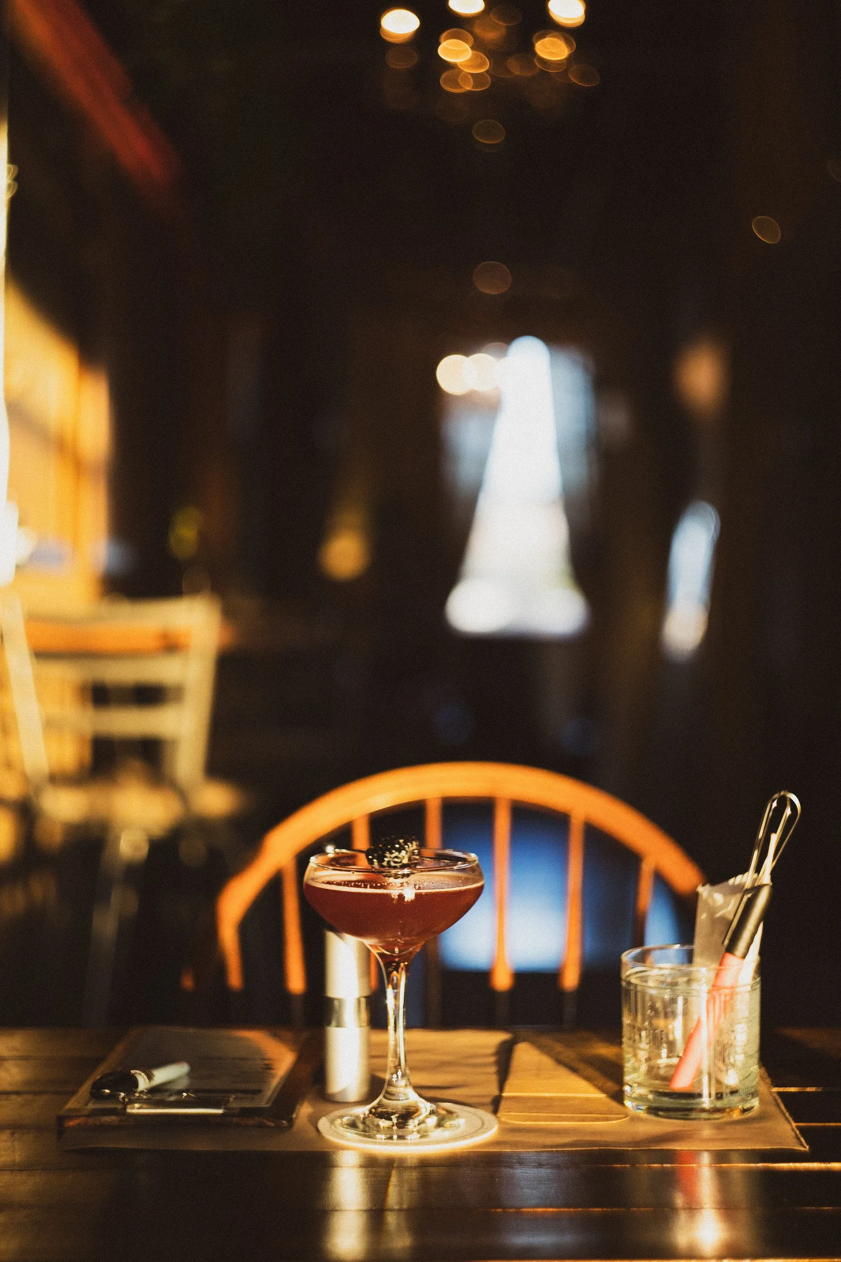A cocktail on a wooden table with a dark background and blurred light spots. The drink is in a coupe glass garnished with blackberries. Nearby, there is a glass with utensils and a scent selection sheet.
