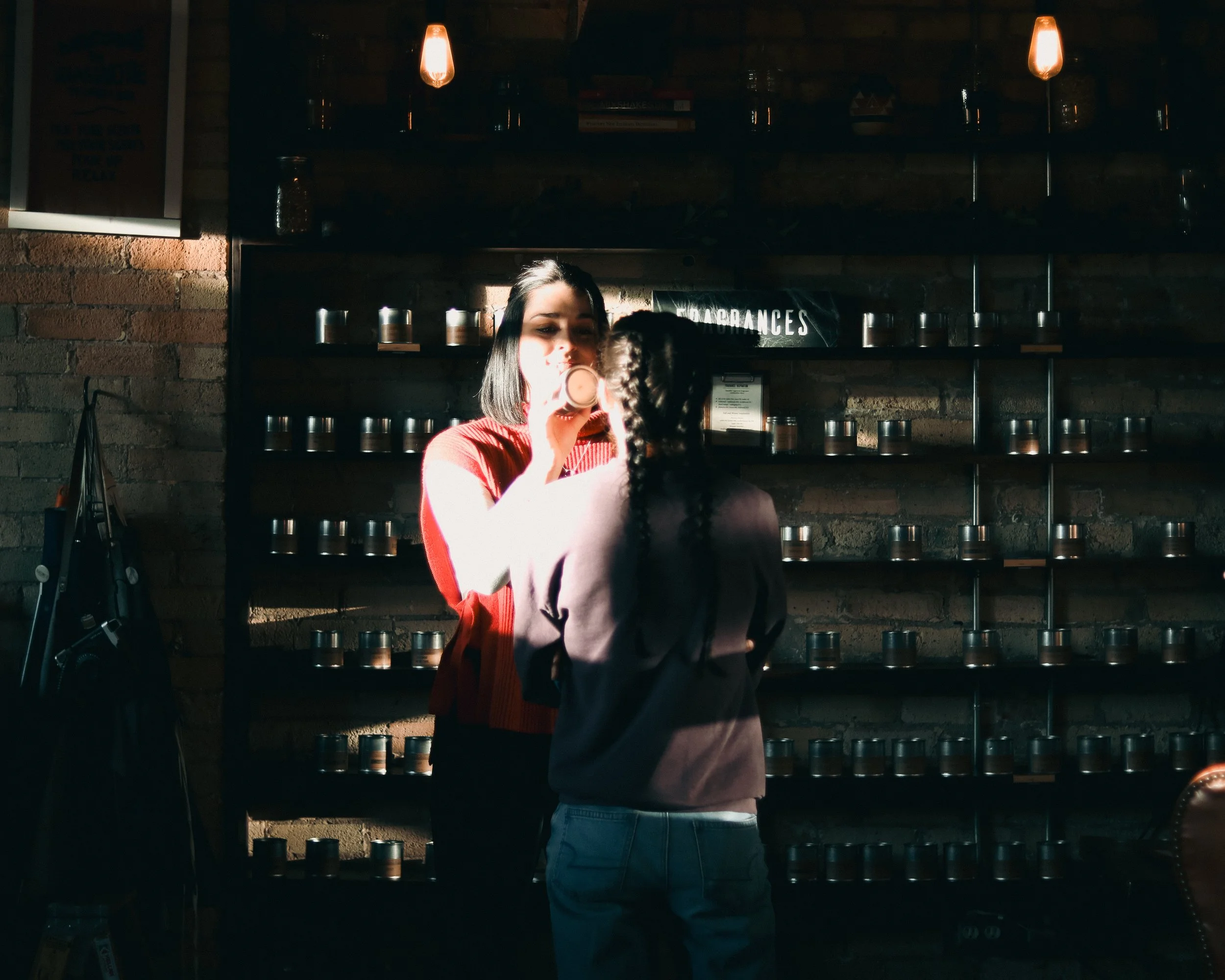 A woman with dark hair in a red top holds a candle to her nose as she interacts with a person with braided hair in a gray top at the candle bar with the fragrance wall set behind them with small metal containers.