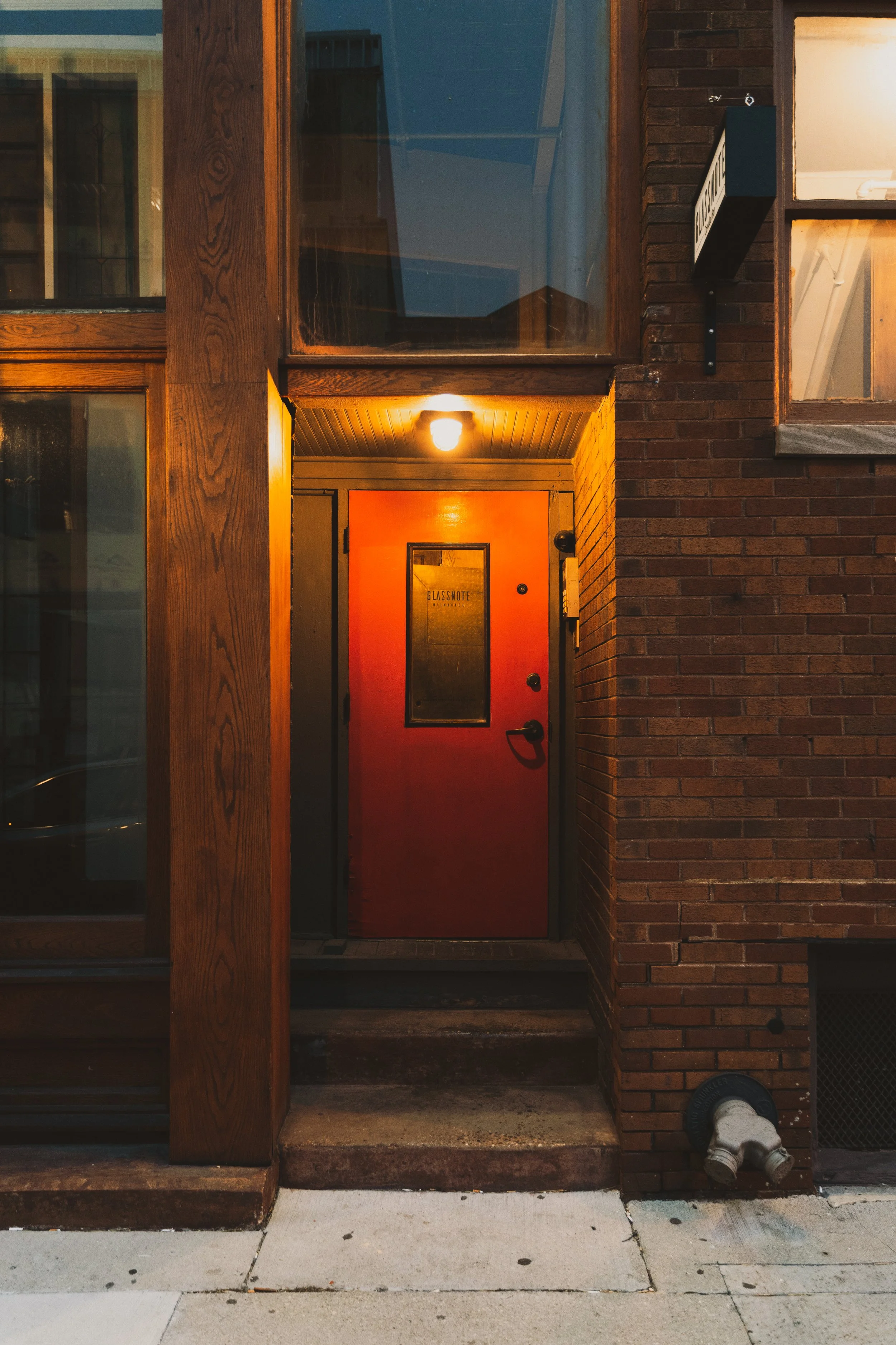 A red door with a window, marked 'GLASSNOTE', located at the entrance of a brick building with a small concrete step and a light overhead, seen from the sidewalk.