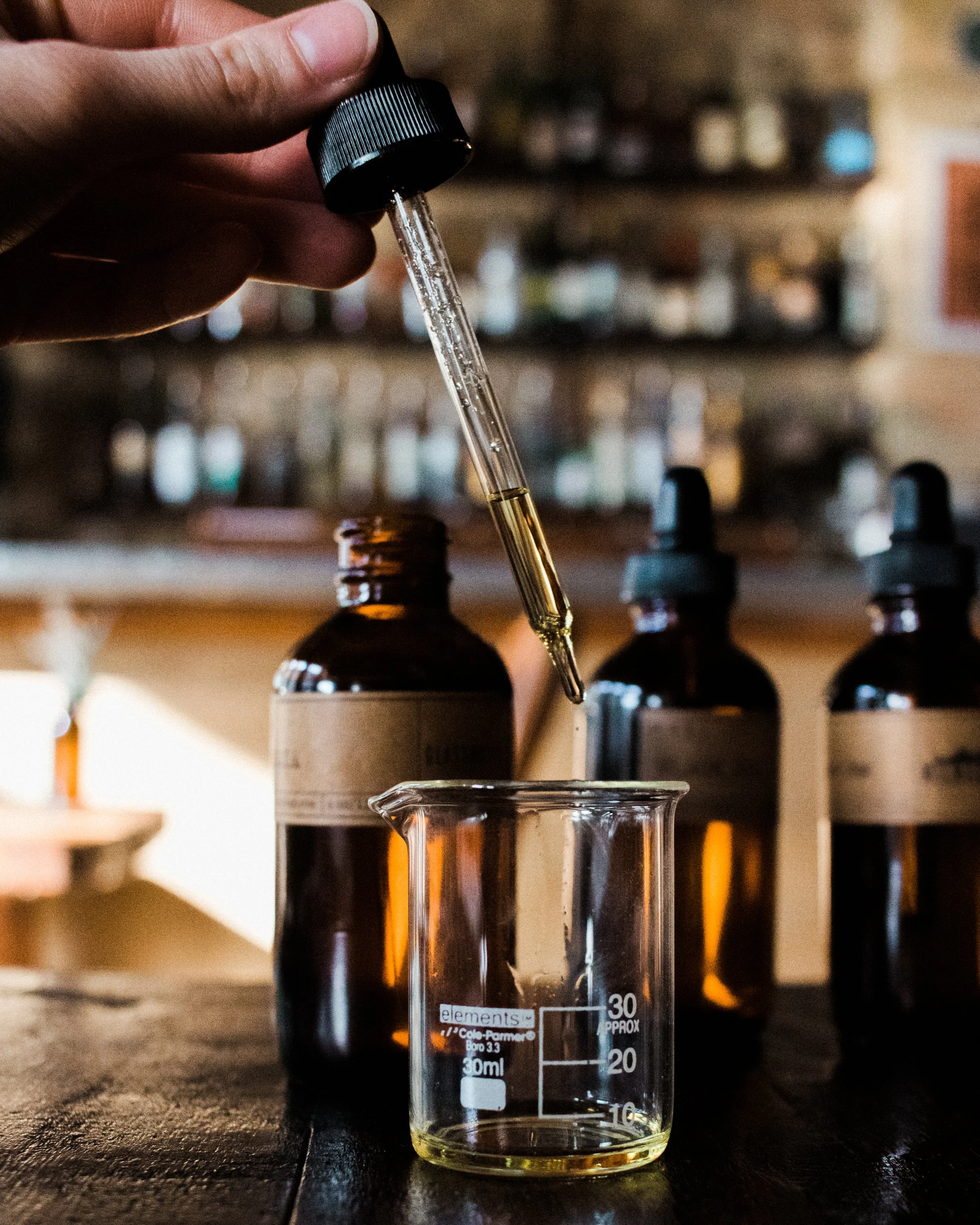 A person holding a glass dropper with a fragrance oil, dropping it into a small glass beaker on a wooden surface, with brown bottles in the background, blending their custom fragrance at the candle bar.