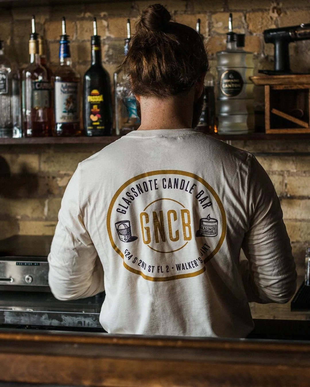 Person with long hair in a bun, wearing a white shirt with a logo that says 'Glassnote Candle Bar' and 'GNCB,' standing behind a zinc bar with liquor bottles on a brick wall background.