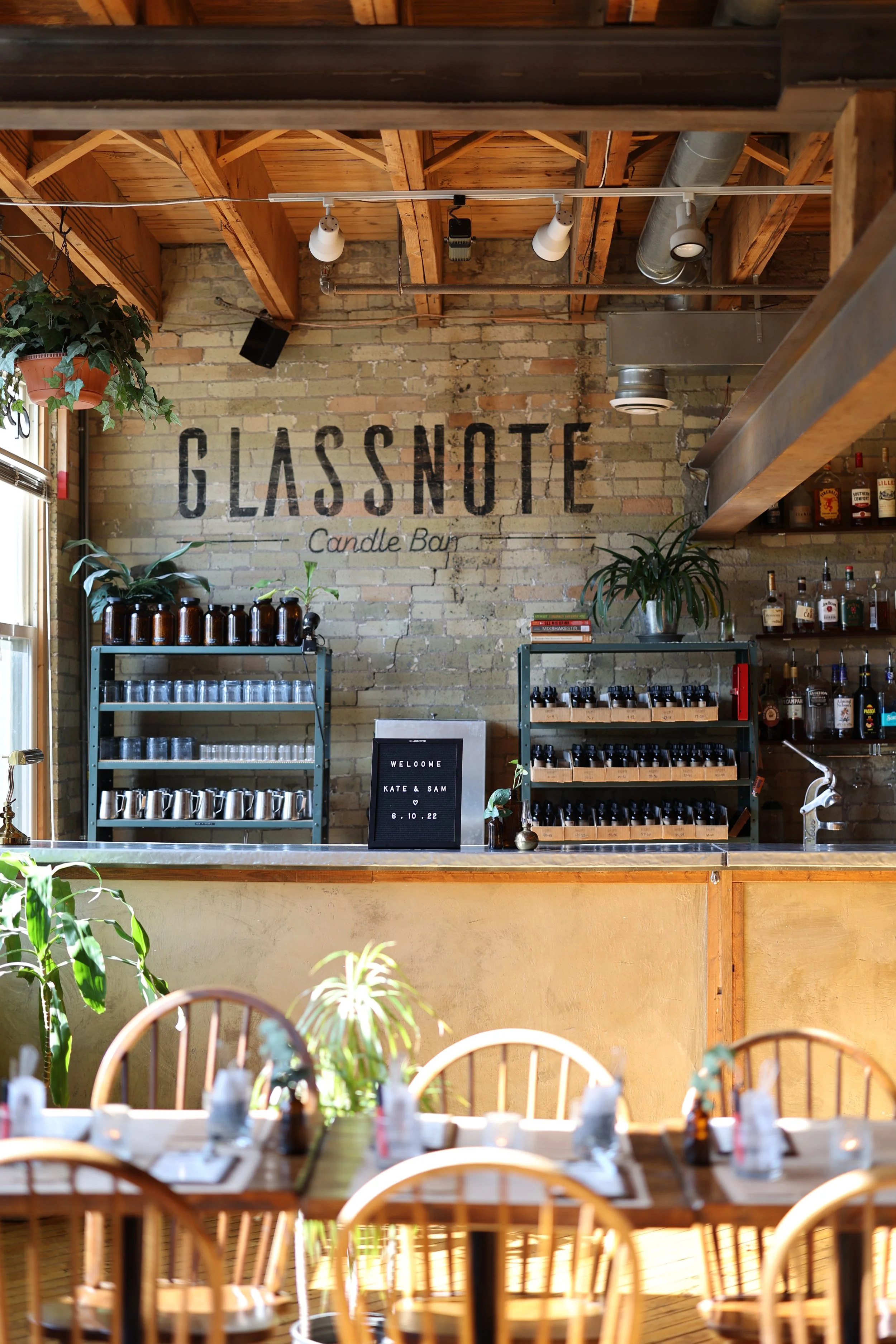 Interior of Glassnote Candle Bar with a brick wall, shelves stocked with candles and bottles, and a table set for the experience with wooden chairs and plants.