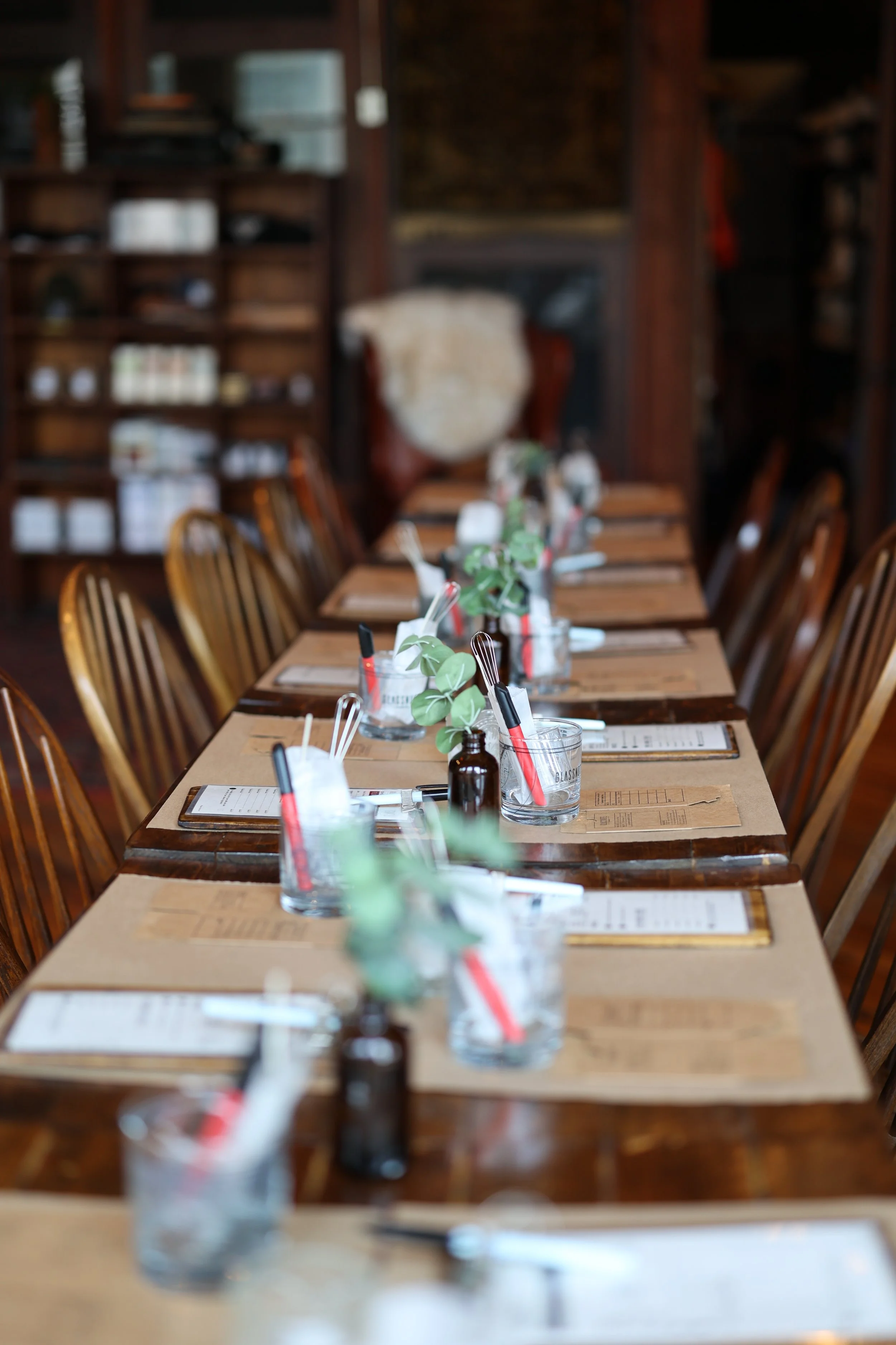 Long dining table set for private event candle making, with wooden chairs, glasses with utensils and fragrance selection cards, in the cozy glassnote candle bar location.