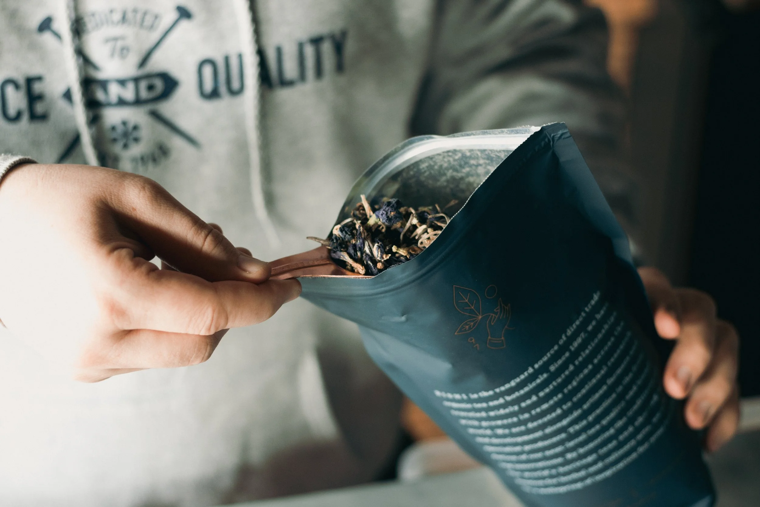 Person opening a resealable bag of dried herbal tea with a wooden spoon inside.