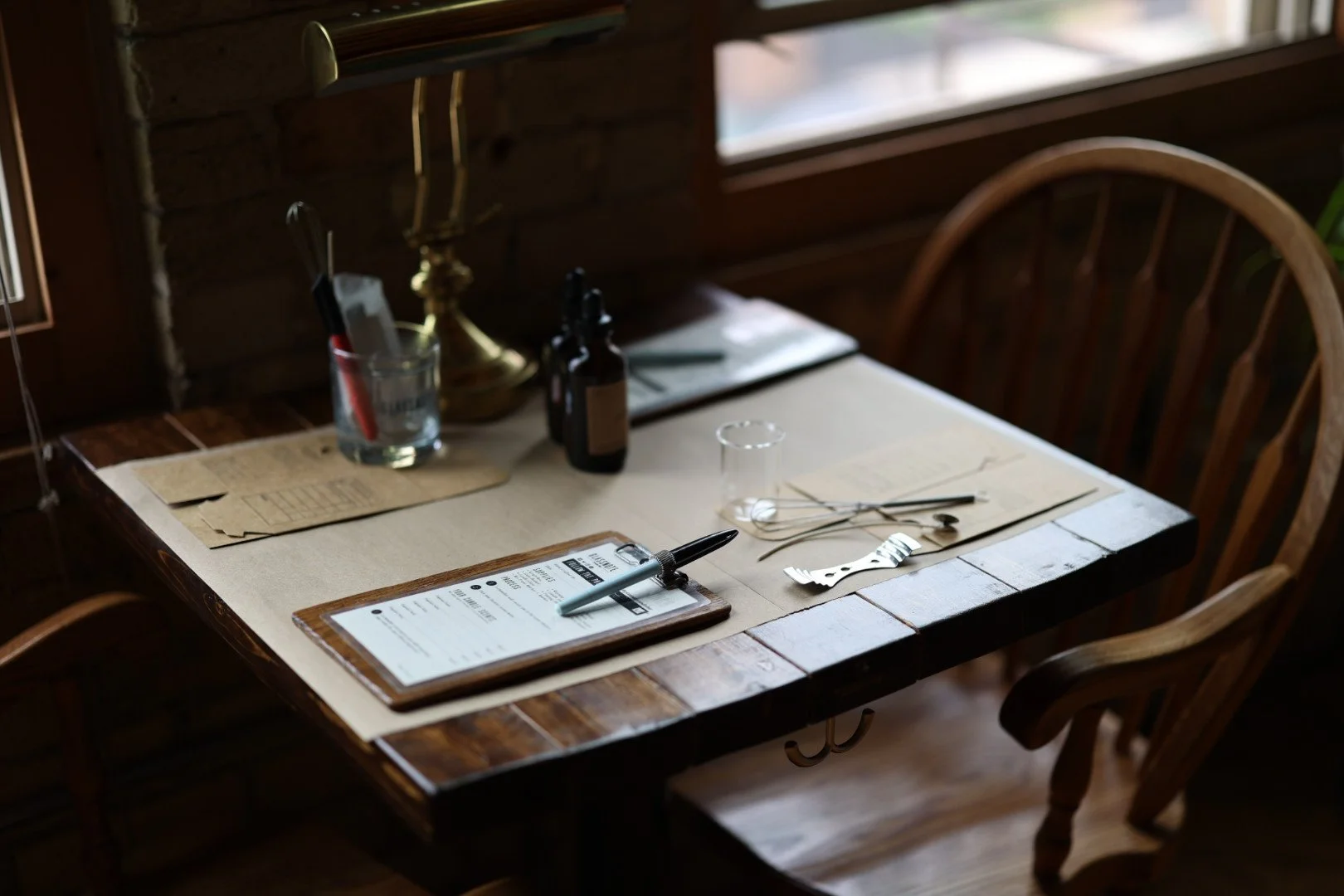 A wooden dining table set for a candle making with handwritten fragrance selection, a pen, and utensils for candle making, near a window with natural light.