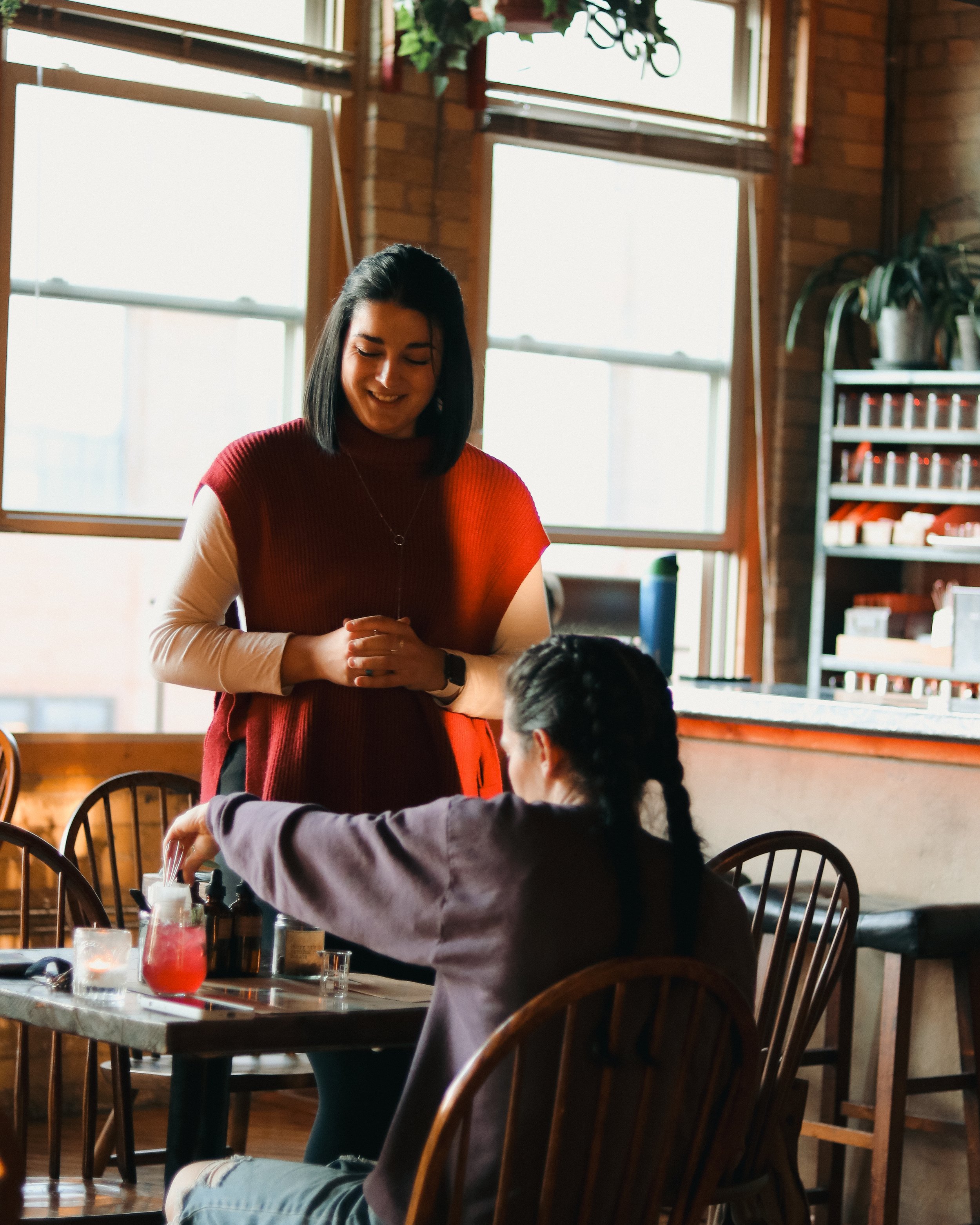 A woman with dark hair and a red sweater standing and smiling inside glassnote candle bar, talking to a girl with braids sitting at a table with drinks and candle making tools. 