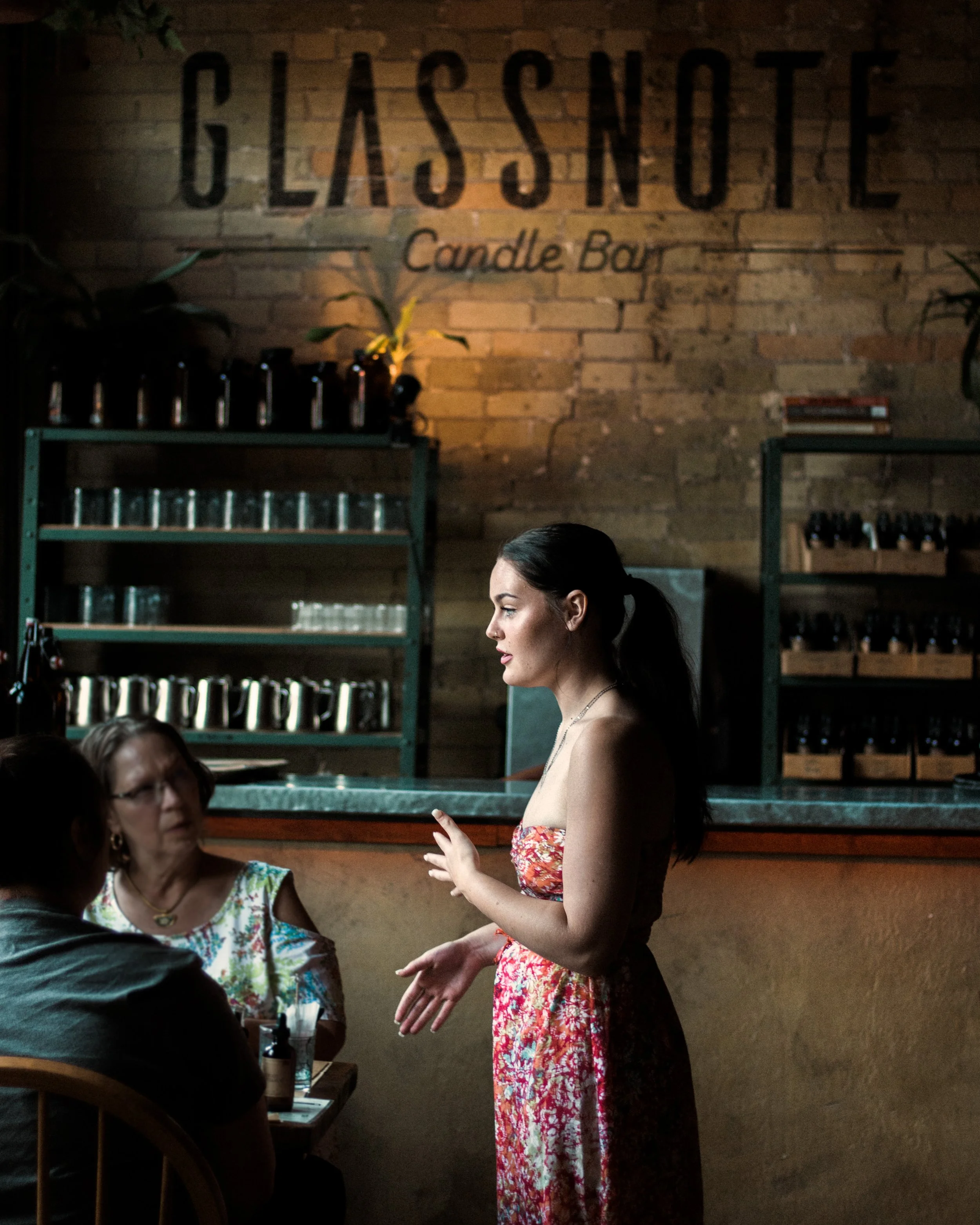 A woman in a floral dress is speaking to a group at the candle bar giving instructions with a brick wall that has the sign 'GLASSNOTE Candle Bar' behind her.