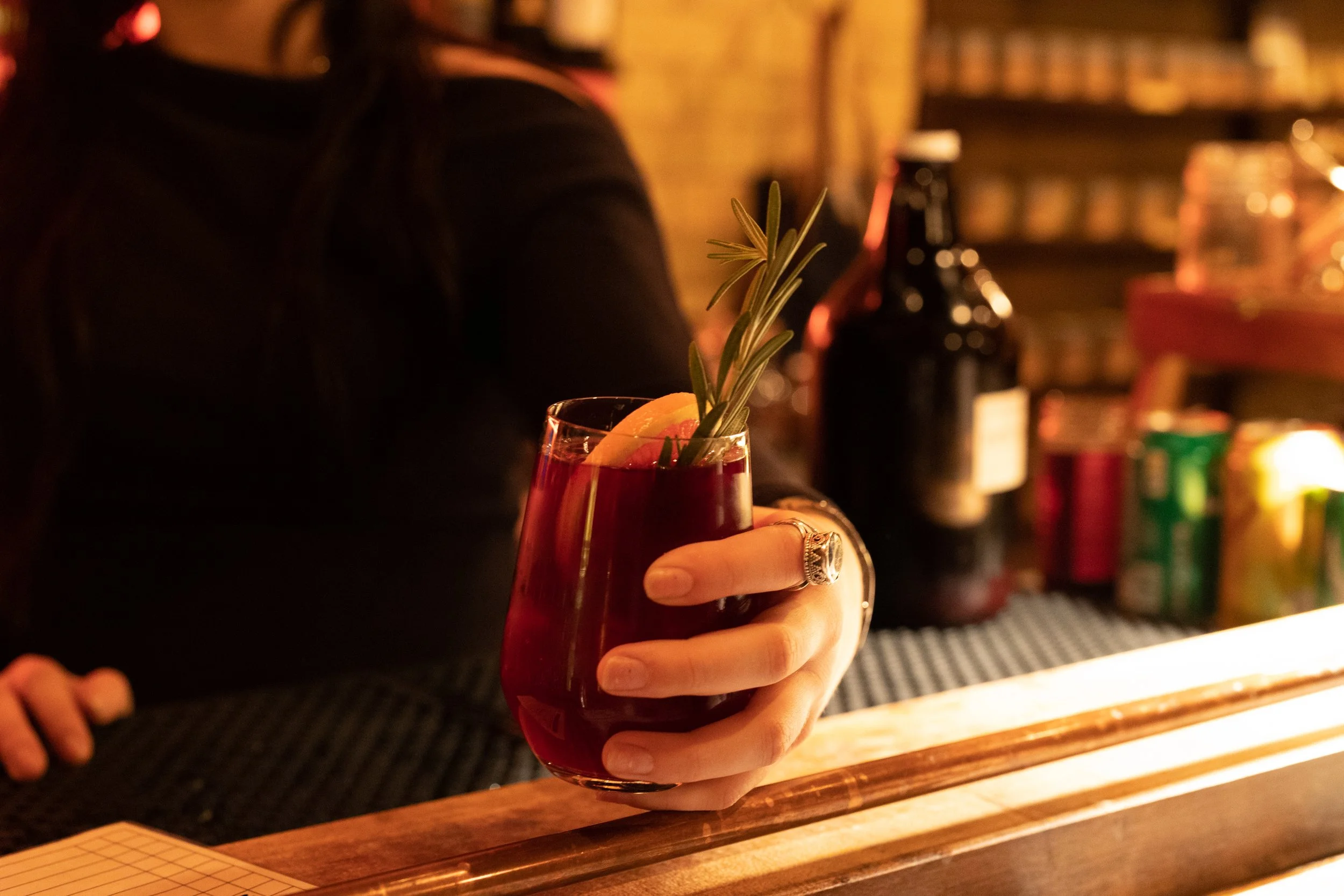 A person holding a glass of red cocktail garnished with a lemon wedge and rosemary sprig at the zinc bar top.