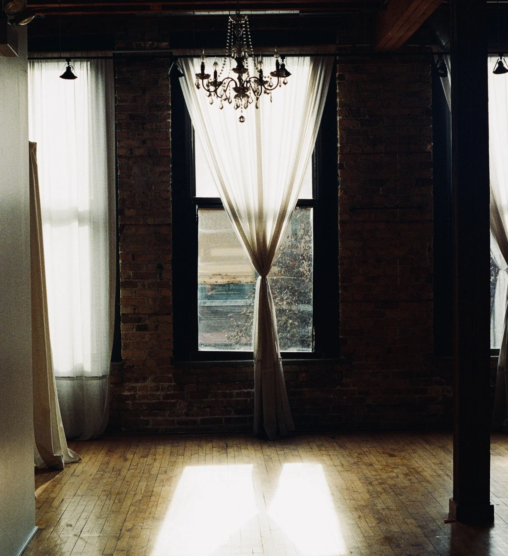 Sunlight streaming through sheer white curtains in a brick-walled room with hardwood floors, chandelier hanging from the ceiling, and large windows with black framing.