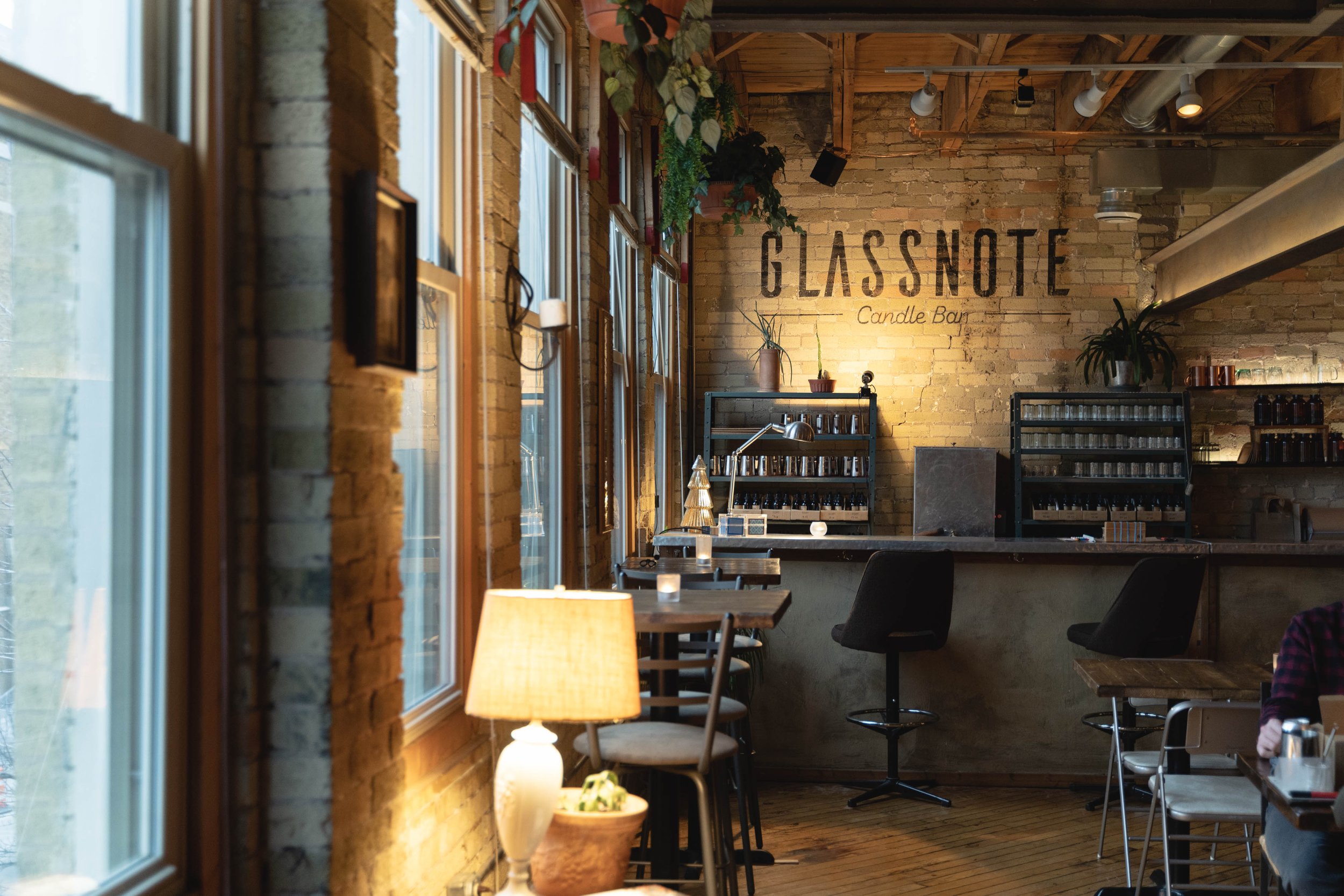 Interior of the candle bar with brick walls, large windows, and warm lighting. The sign on the wall reads "Glassnote Candle Bar." There are tables with chairs, shelves with candles, and decorative plants.