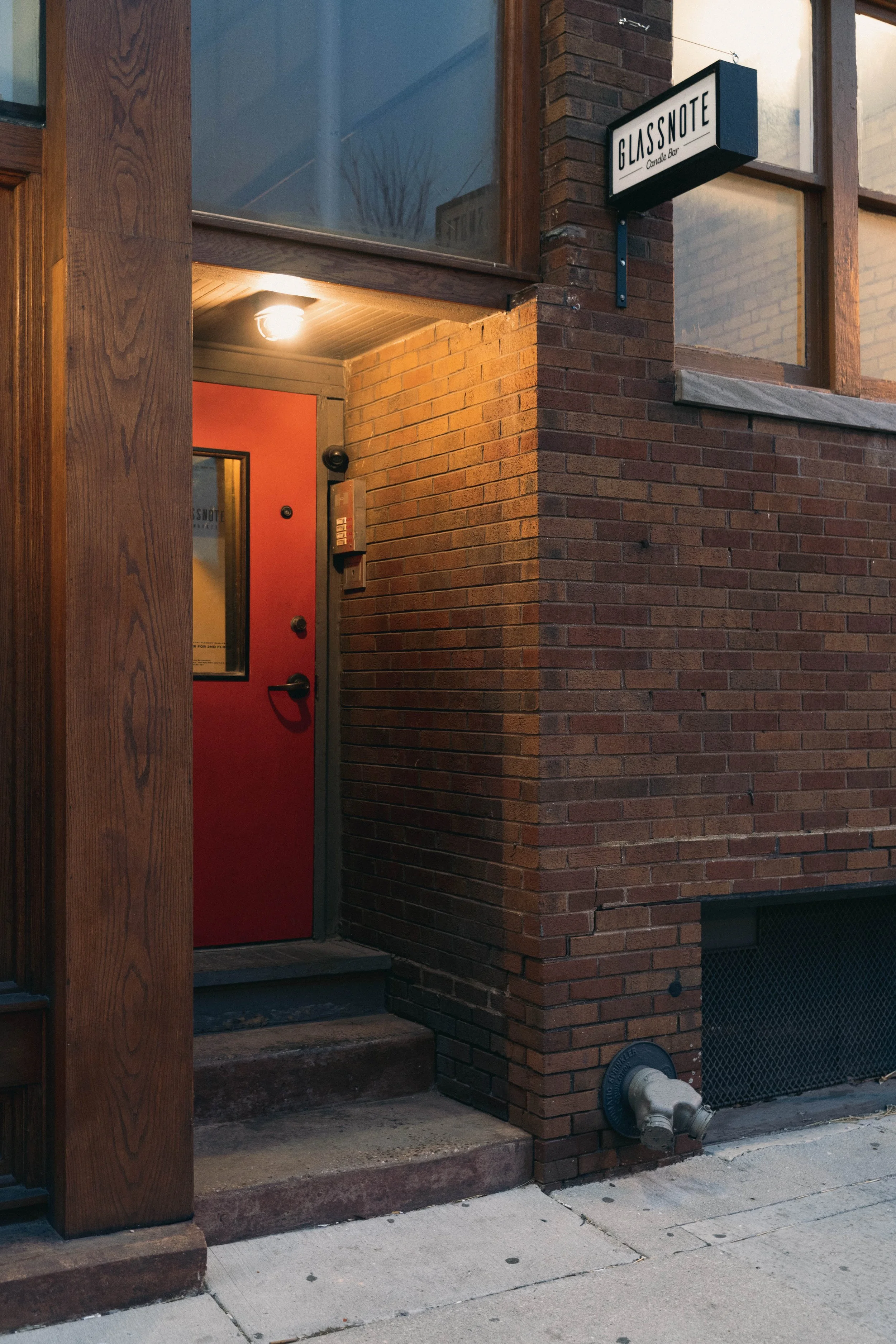 Brick building entrance with a red door, a sign that reads 'Glassnote Candle Bar,' and a warm light overhead. A view from the sidewalk.