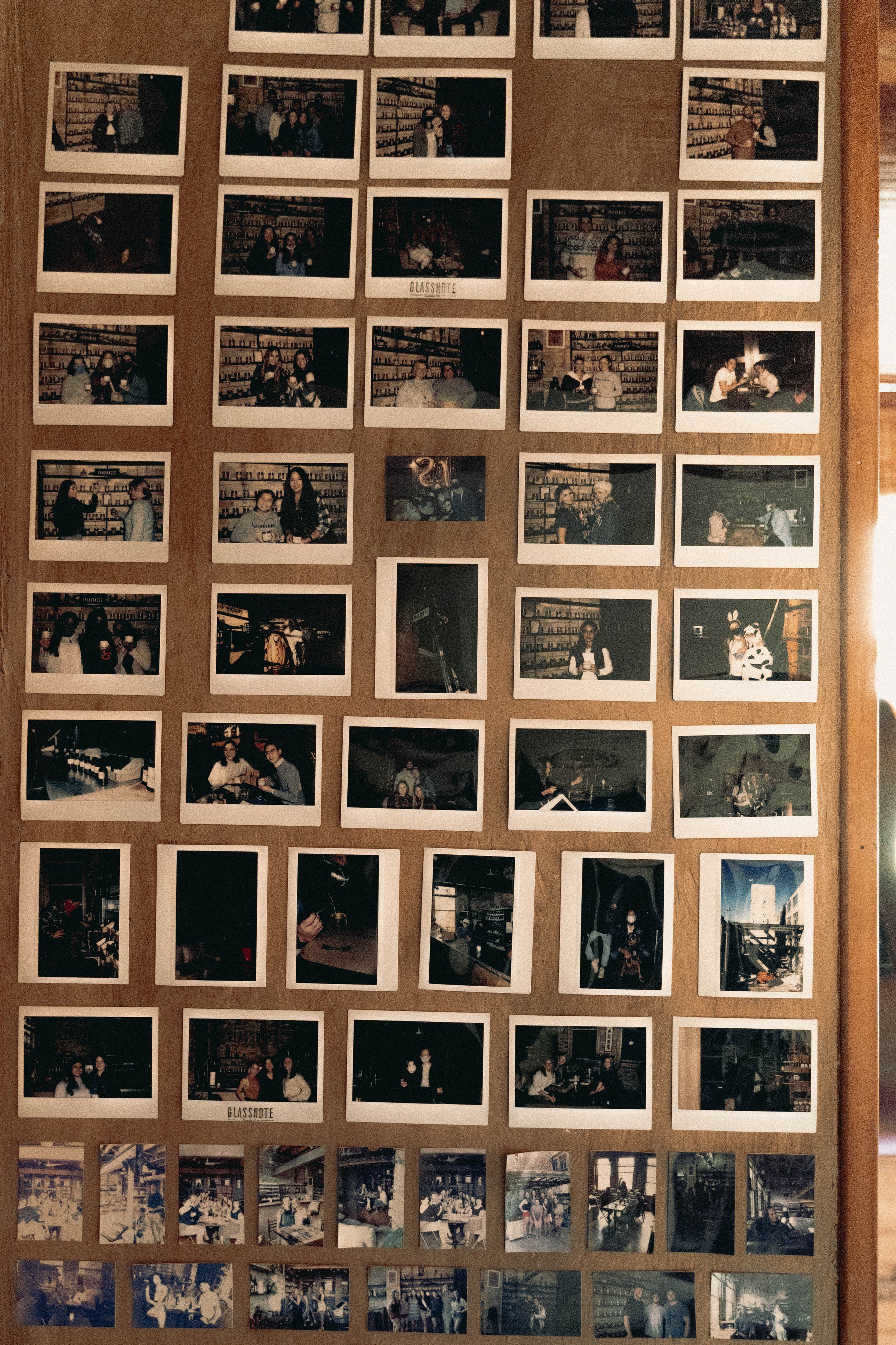 A wall decorated with many polaroid photos of people gathered at glassnote candle bar, some taking group photos, and some sitting alone or with others, with a wooden wall background.