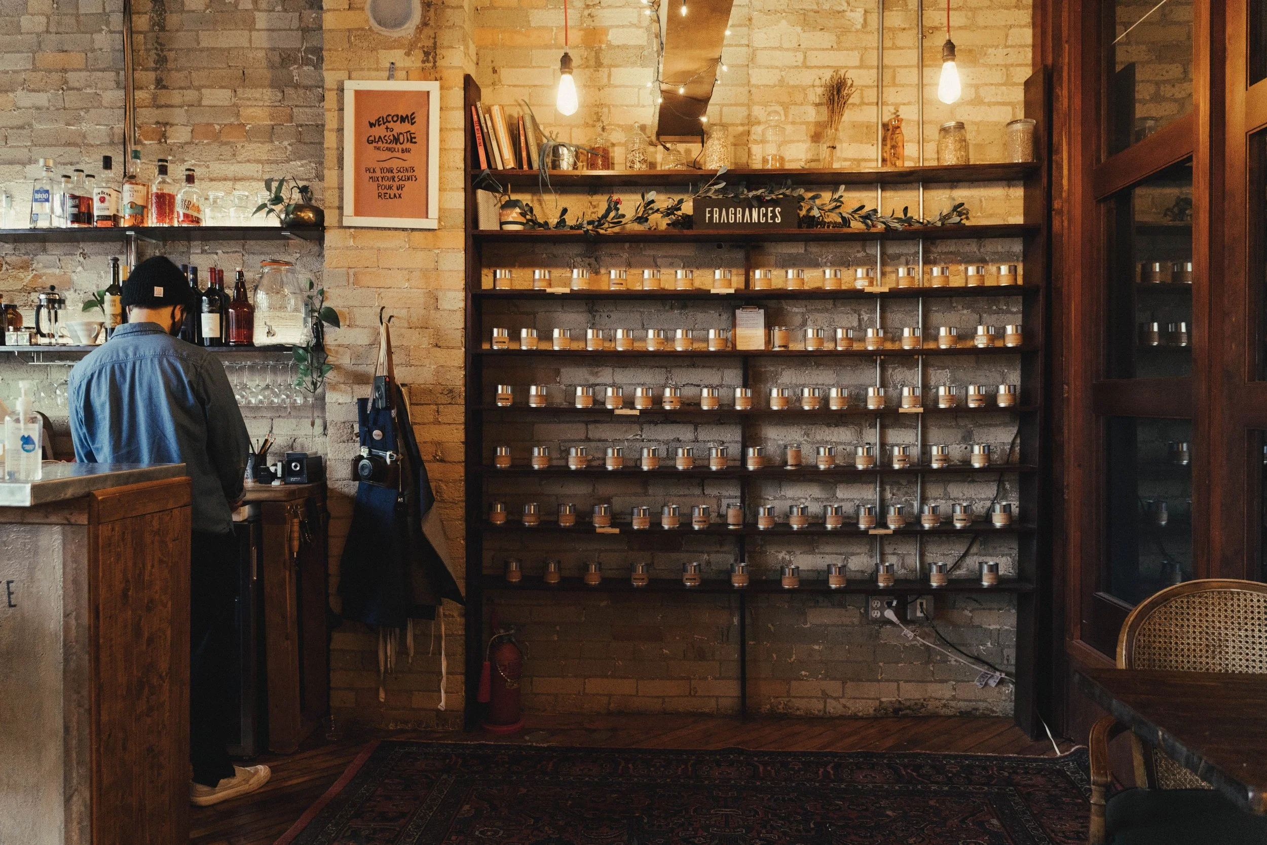 Interior of the candle bar with a brick wall featuring the fragrance wall, shelf labeled 'Fragrances' filled with small candle jars. To the left, a person is working behind a counter with bottles and glasses.