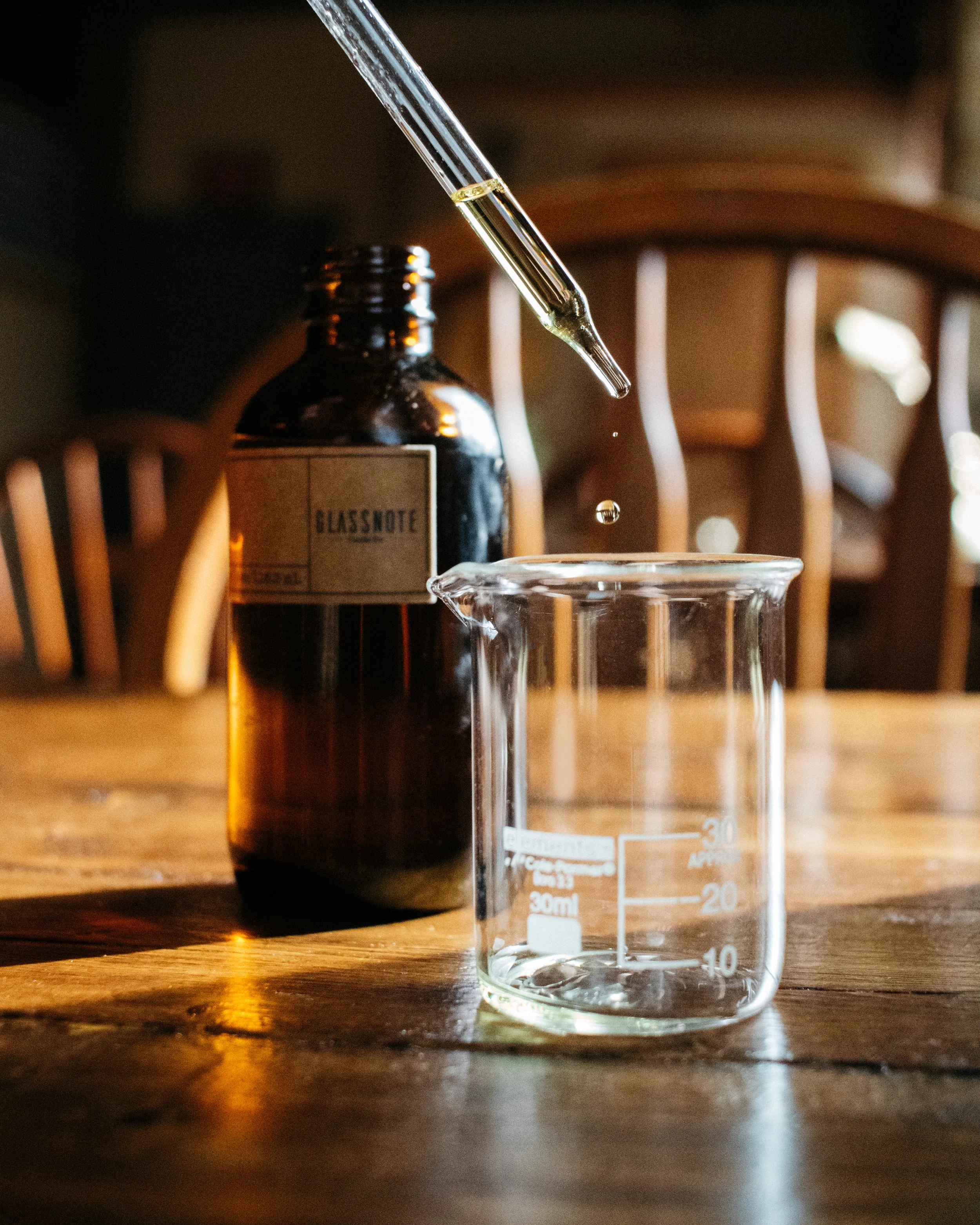 A glass dropper above a brown glass bottle labeled 'Glass Note' with a pipette releasing a droplet of fragrance oil into a small glass beaker on a wooden table.