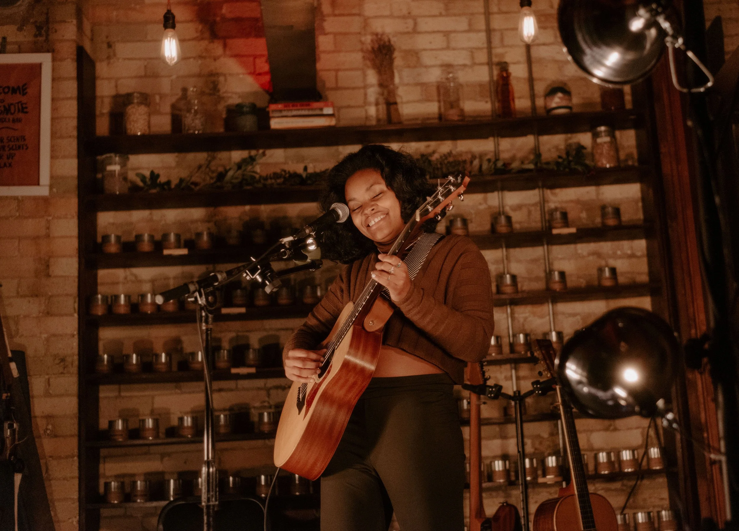 A woman playing an acoustic guitar and singing into a microphone on stage in front of the fragrance wall, smiling with eyes closed, in a cozy, warmly lit brick-walled venue.