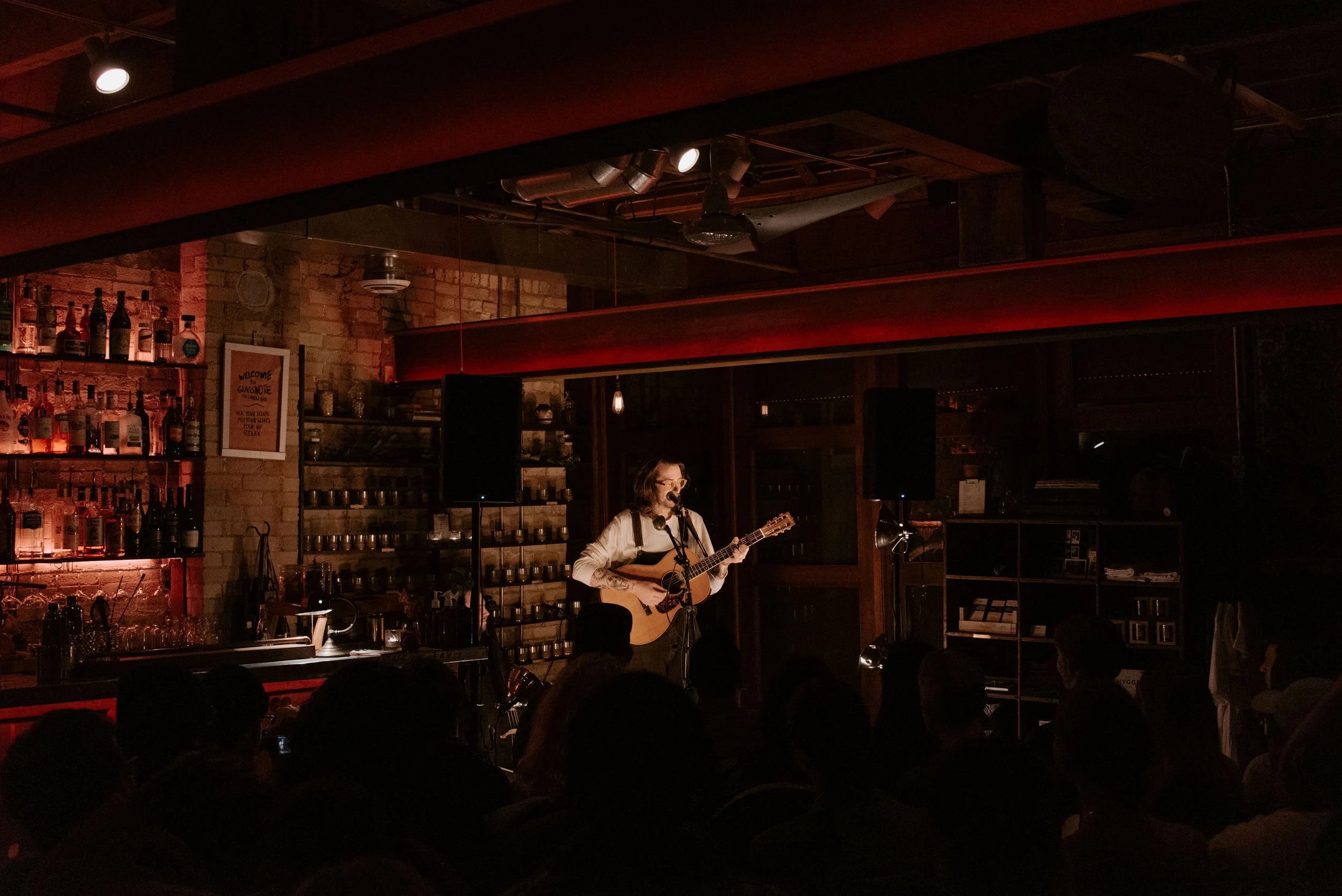 A person with glasses playing an acoustic guitar and singing into a microphone on a small stage in the dimly lit candle bar, with an audience watching.