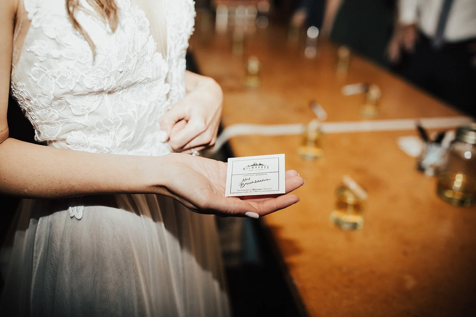 A woman in a white lace wedding dress holds a small square card with the text 'Milwaukee' and 'the Brewers' in front of a wooden table with small glasses.