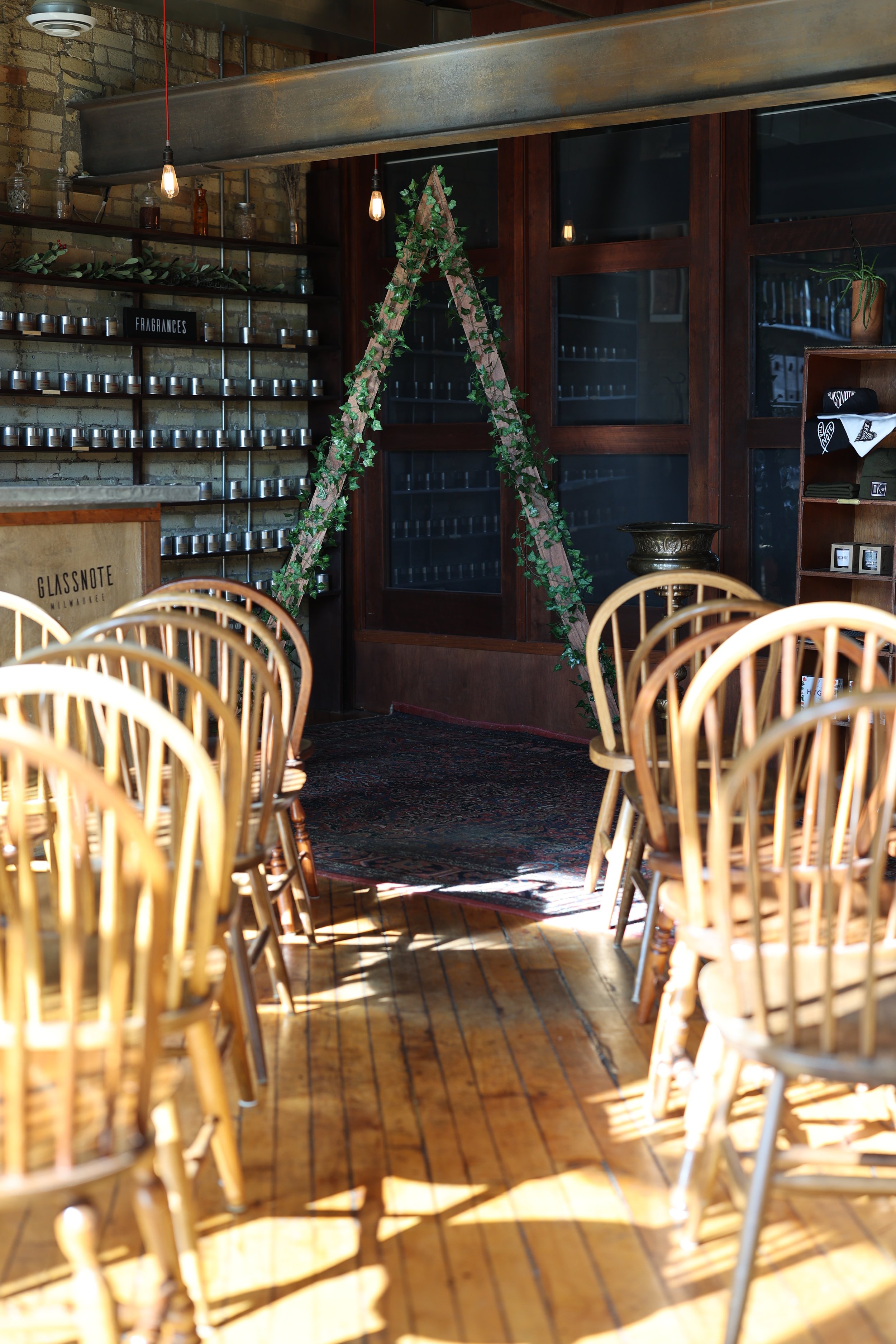Inside the candle bar with wooden chairs and tables, brick wall with shelves of candles and a sign that says 'FRAGRANCES', a decorative triangle frame with greenery, a wooden counter, and large windows for a wedding.