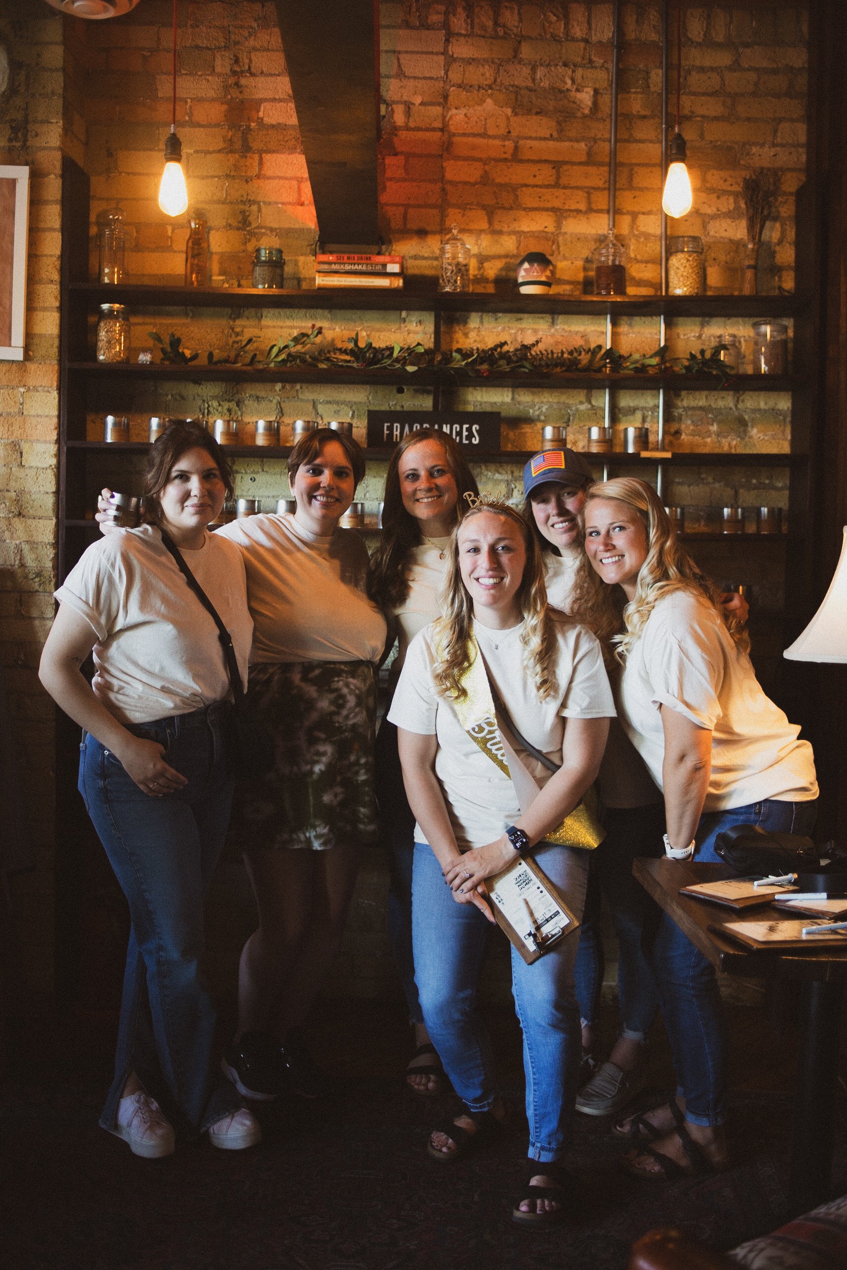 Group of six women smiling and posing in a cozy, warmly lit candle bar with a brick wall backdrop, shelves with jars and books, and hanging lights.
