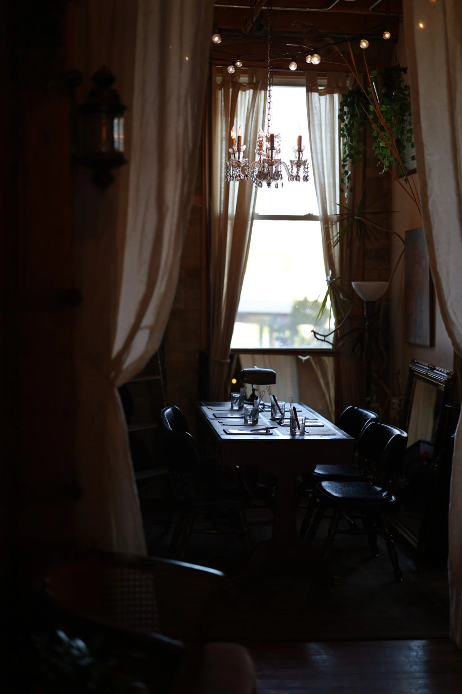 A dimly lit hallway with a table set with glasses and candle making materials. Large window with light curtains, chandelier, and string lights hang from the ceiling. Curtains frame the opening, revealing sunlight outside.