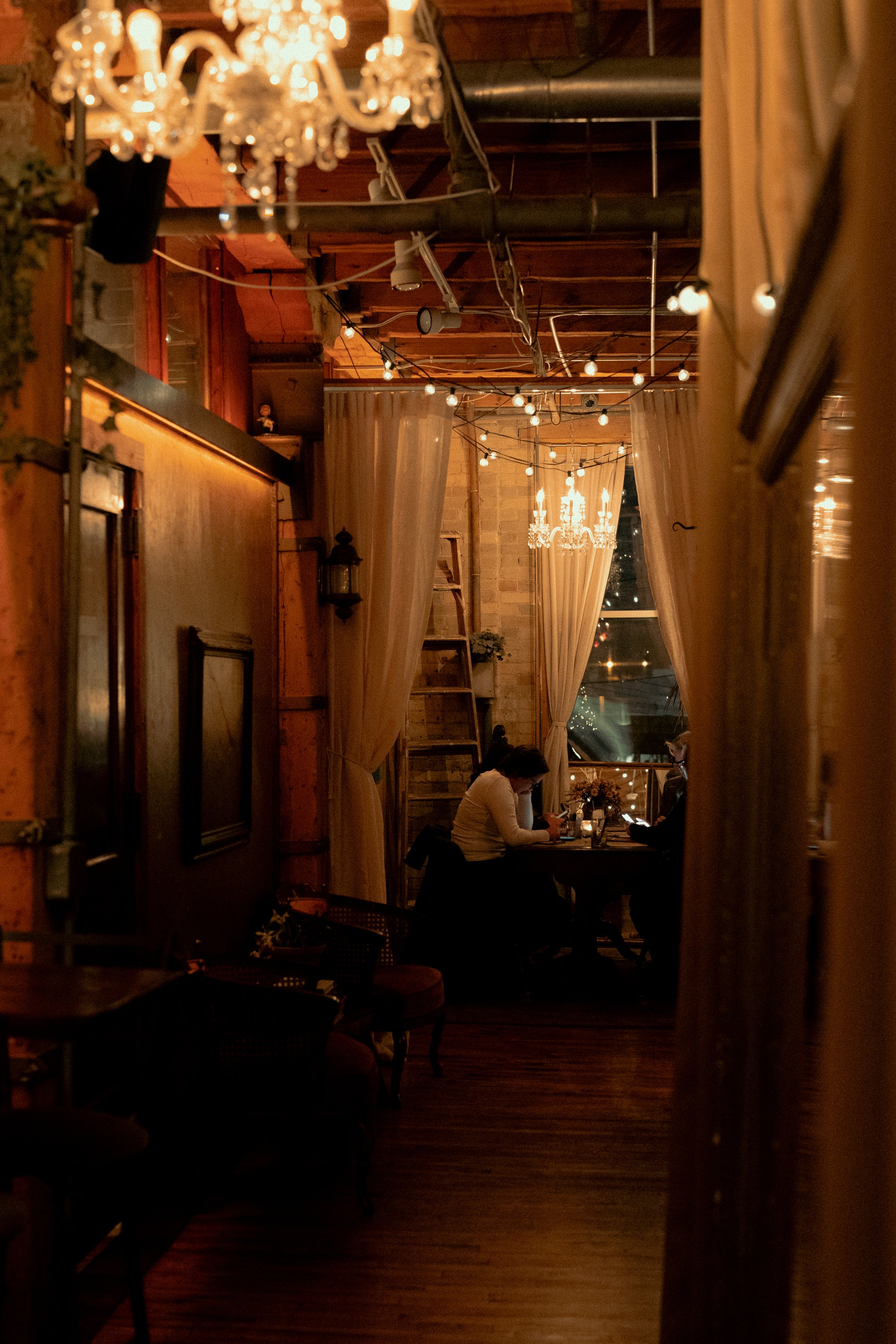 A cozy candle bar interior with dim lighting, chandeliers, string lights, wooden walls, curtains, and two people seated at a table near a window with city lights visible outside.