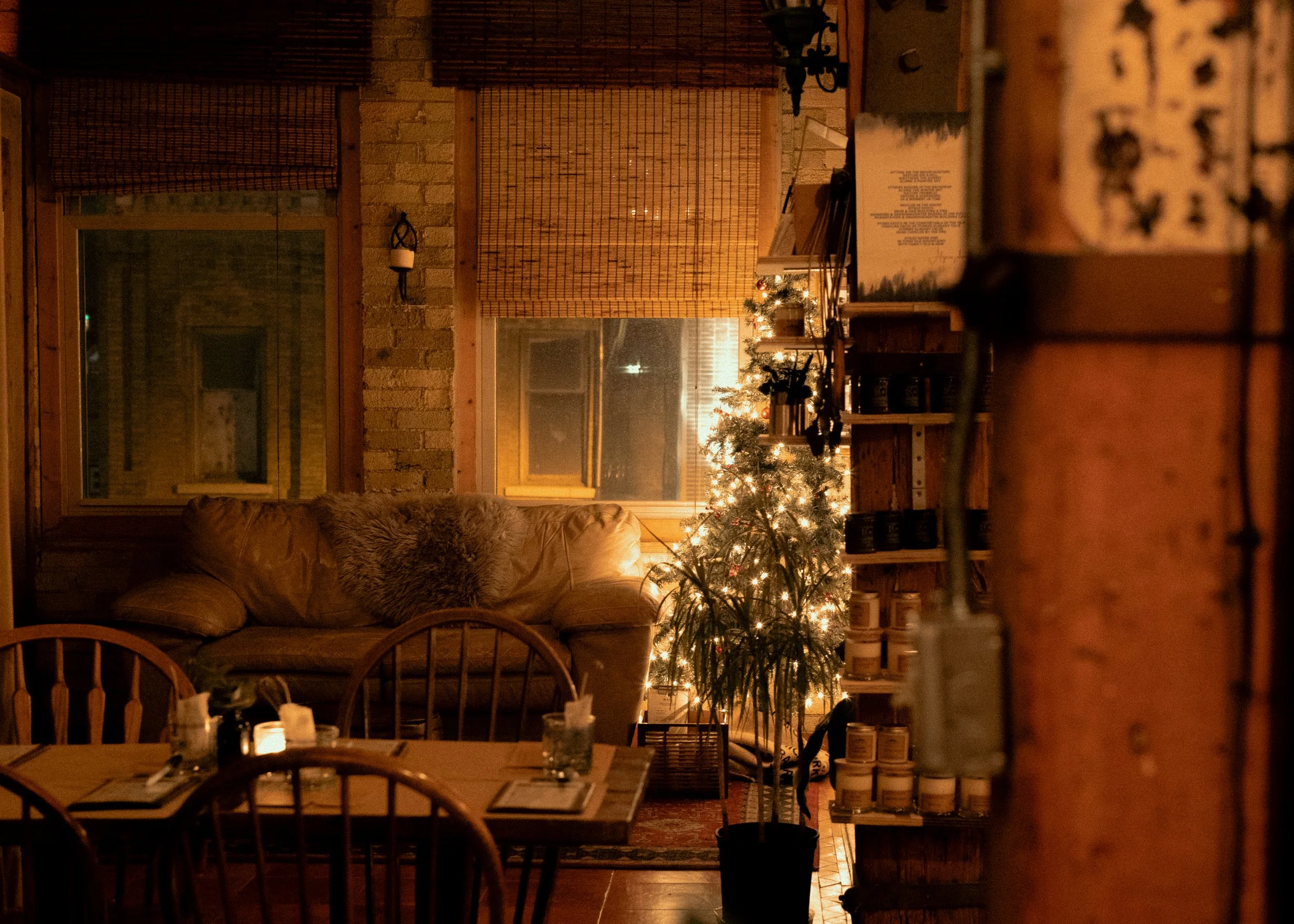 The candle bar during Christmas with a decorated Christmas tree, a leather couch with a furry pillow, wooden dining table with placemats and candles, brick wall, windows, and wooden shelves filled with candles.