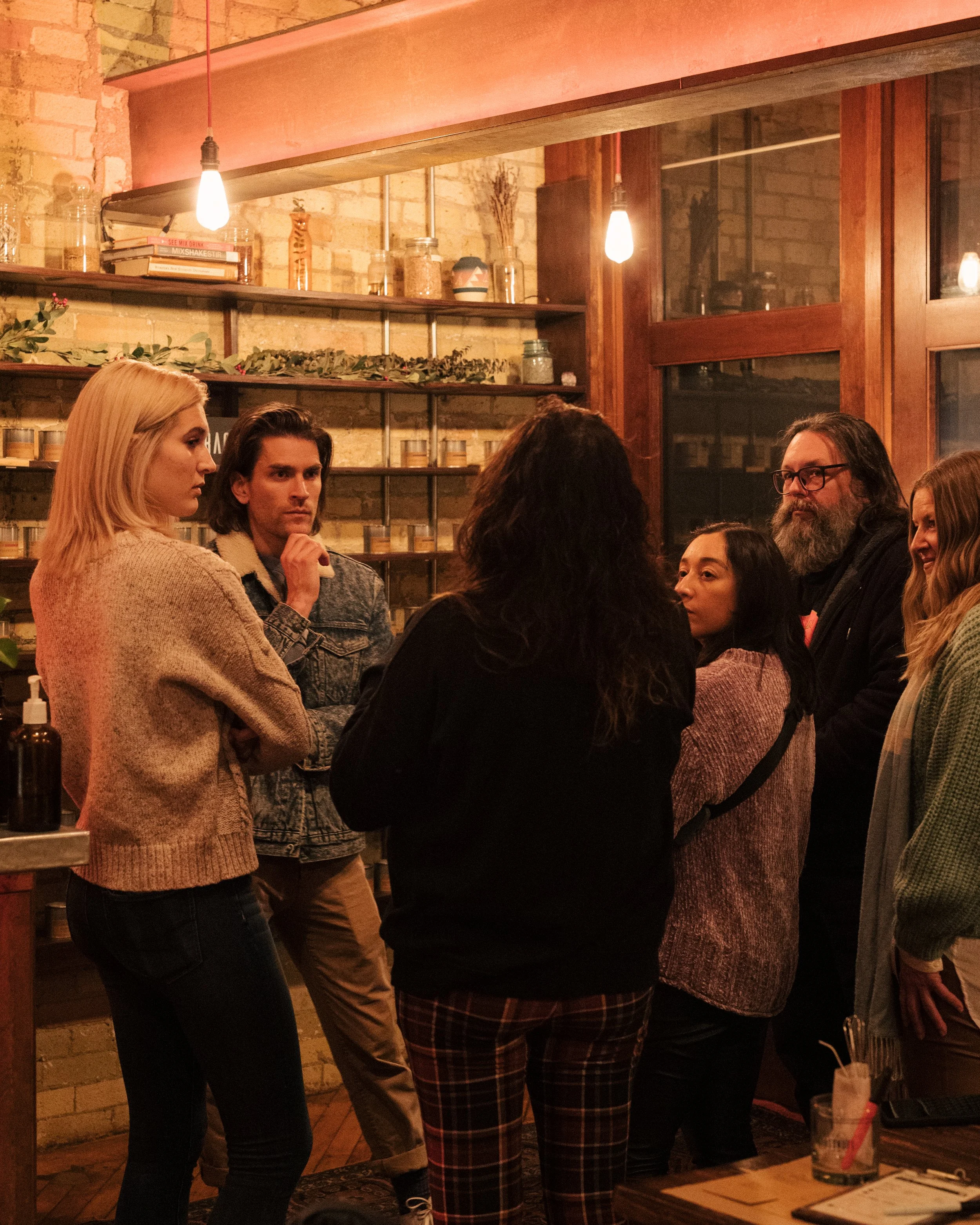 Group of six people conversing in a cozy, warmly lit candle bar in front of the fragrance wall while they select their fragrances.