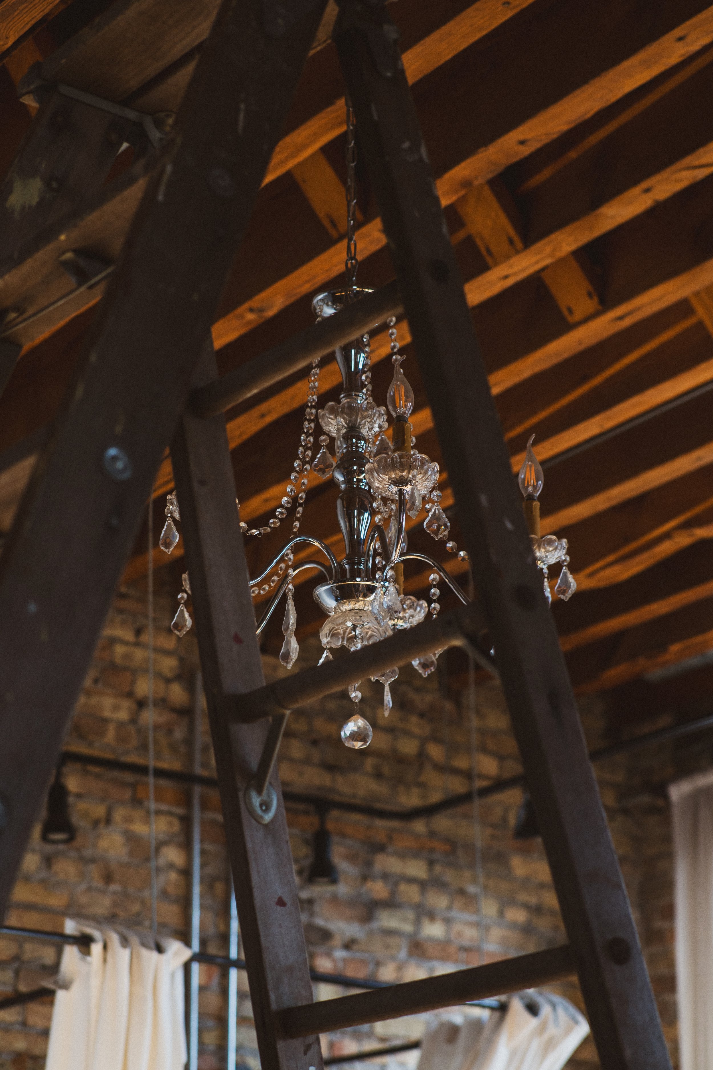 A crystal chandelier hanging from the ceiling, viewed through a ladder in front of a brick wall and wooden beams.