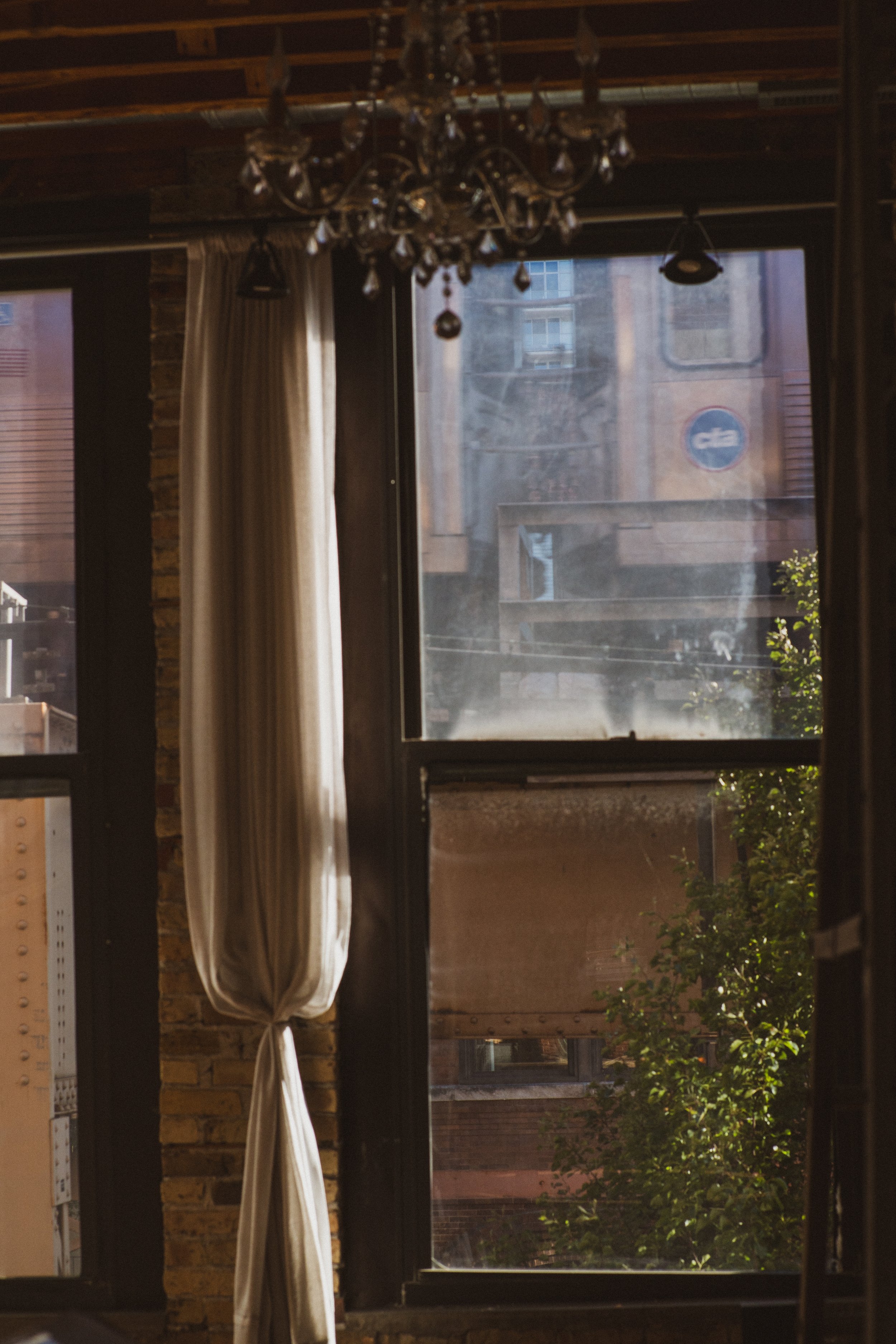 View of city buildings outside a window with partially drawn white curtain and chandelier hanging inside.