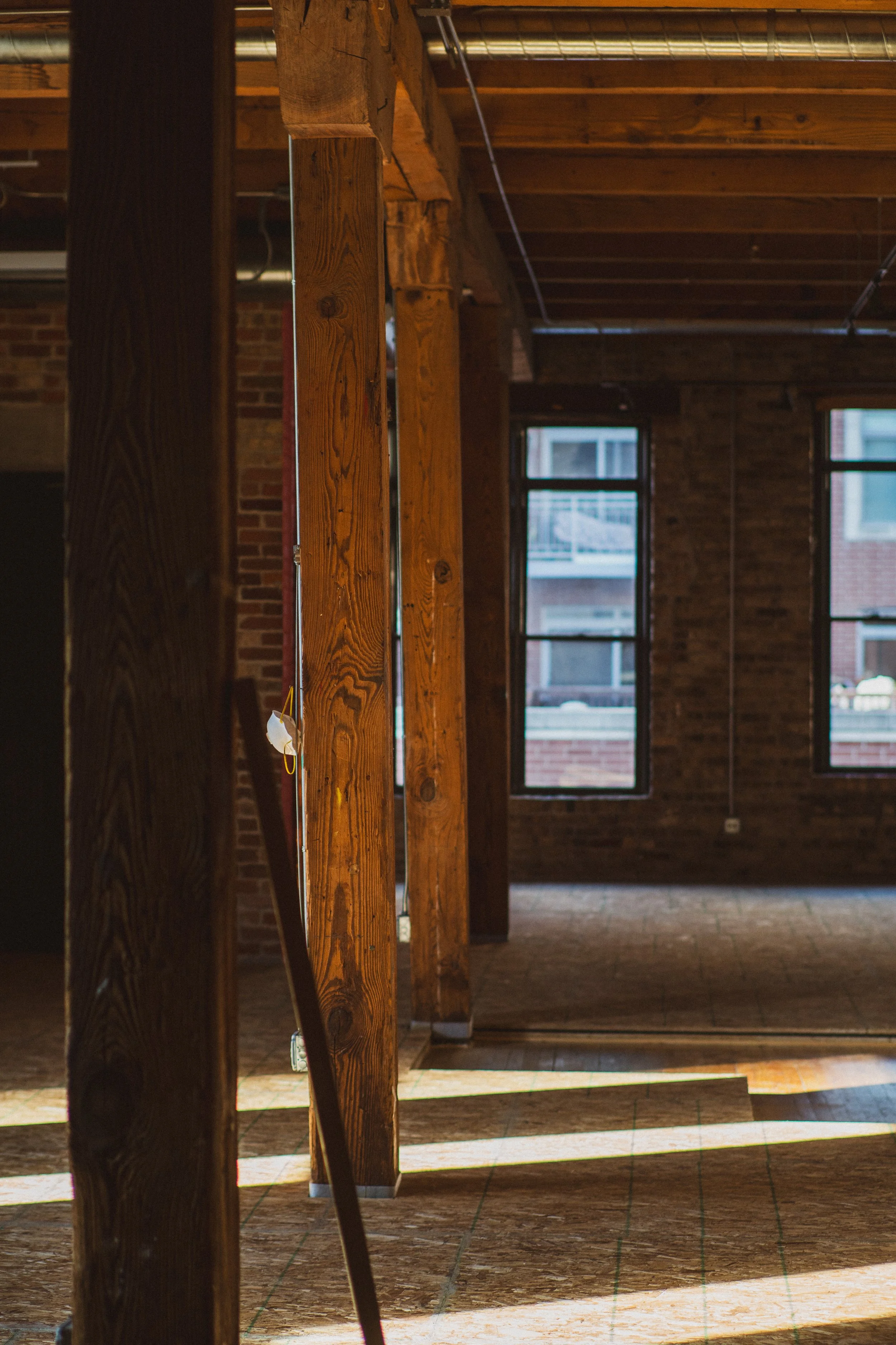 Interior of an unfinished building with exposed wooden beams and brick walls, sunlight streaming through windows onto a wooden floor.