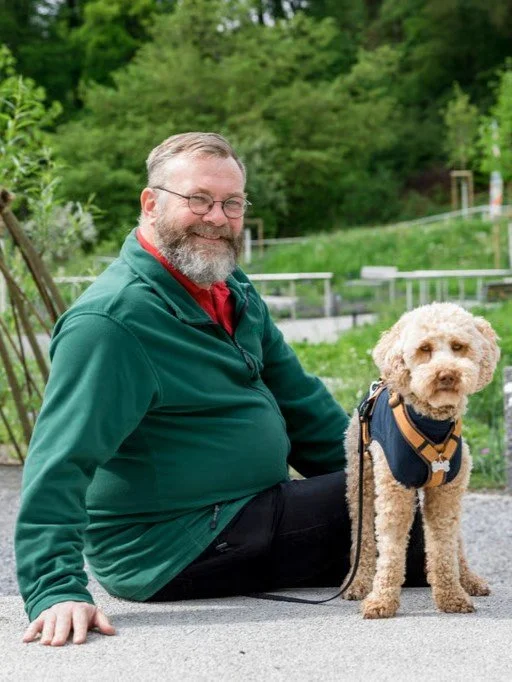 Ein Mann mit Brille und grauem Bart sitzt auf dem Boden, neben einem kleinen, lockigen Hund mit Halsband in einer grünen, natürlichen Umgebung.