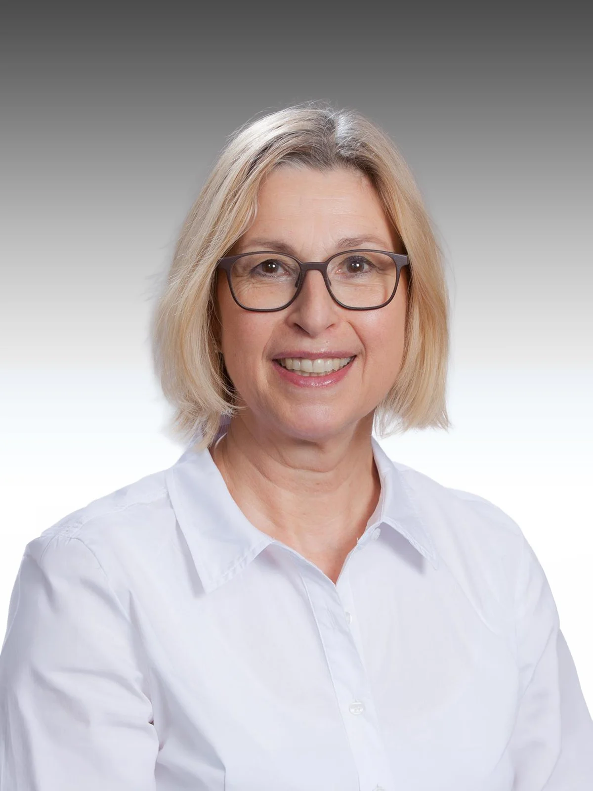 Porträt einer lächelnden Frau mit Brille, kurzen blondem Haar und weißem Hemd vor einem grauen bis weißen background.