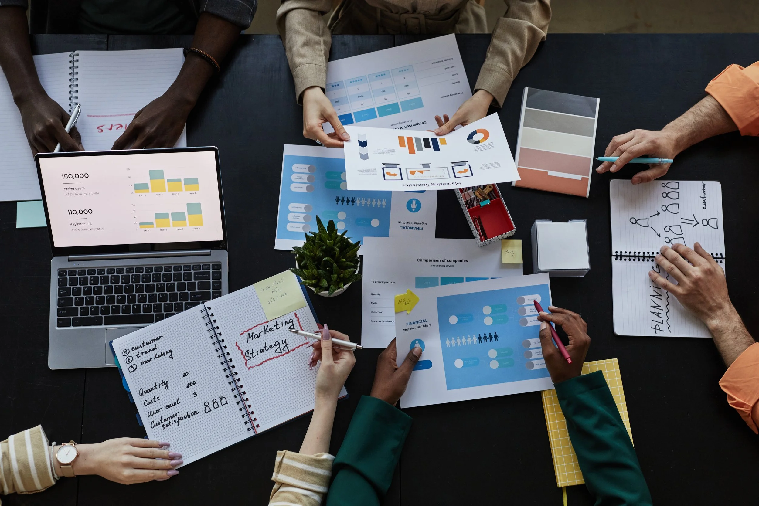 Mehrere Personen arbeiten an einem Tisch mit Notizbüchern, Diagrammen, einem Laptop und Papier, in einer Besprechung oder Teamarbeit.