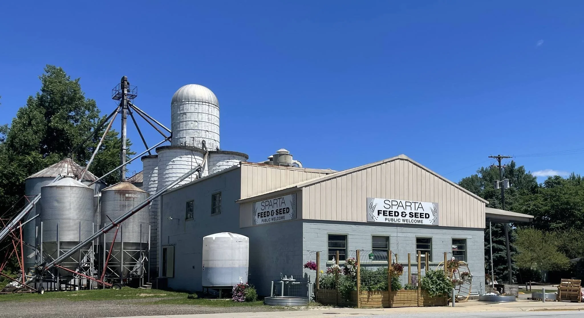A farm building with signs that read 'Sparta Feed & Seed', featuring silos, grain bins, and a clear blue sky.