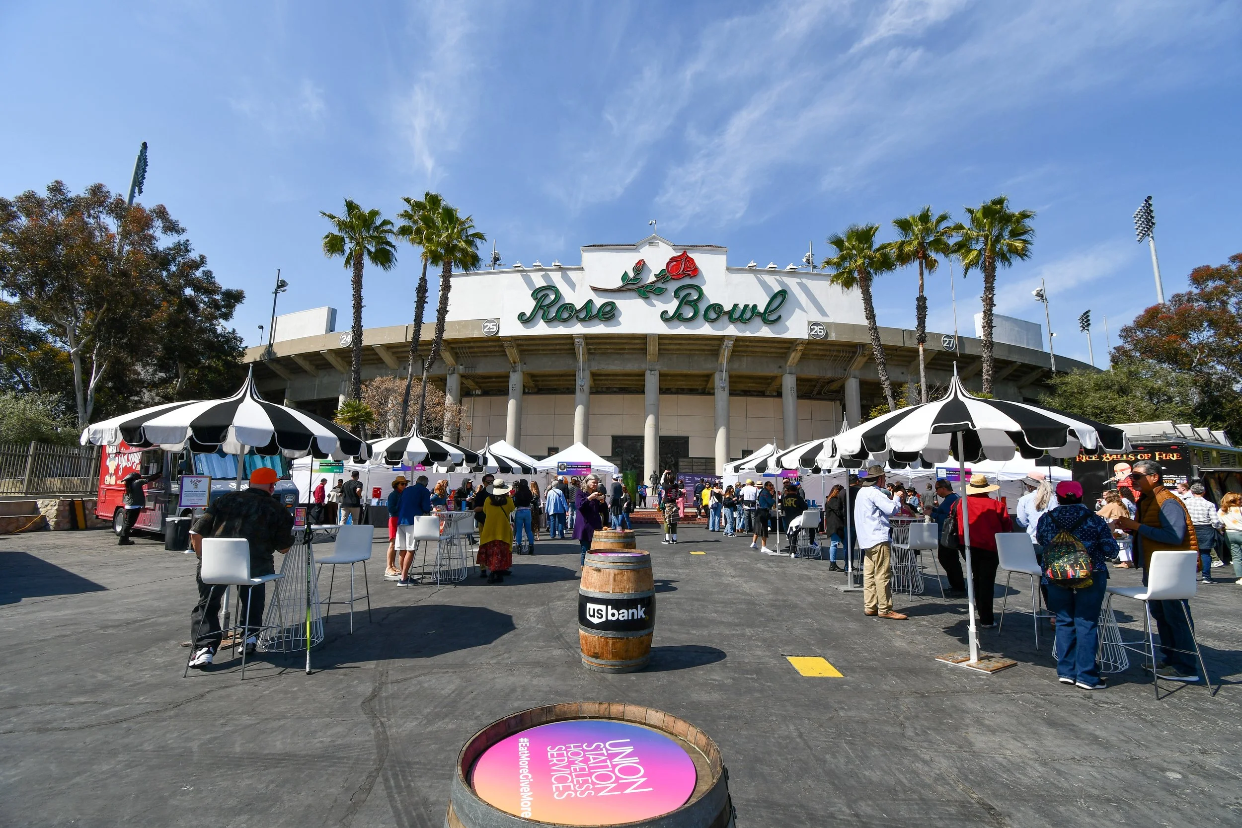 People lining up outside the Rose Bowl stadium for Masters of Taste, with black and white striped umbrellas and a barrel labeled US Bank in the foreground, under a blue sky.