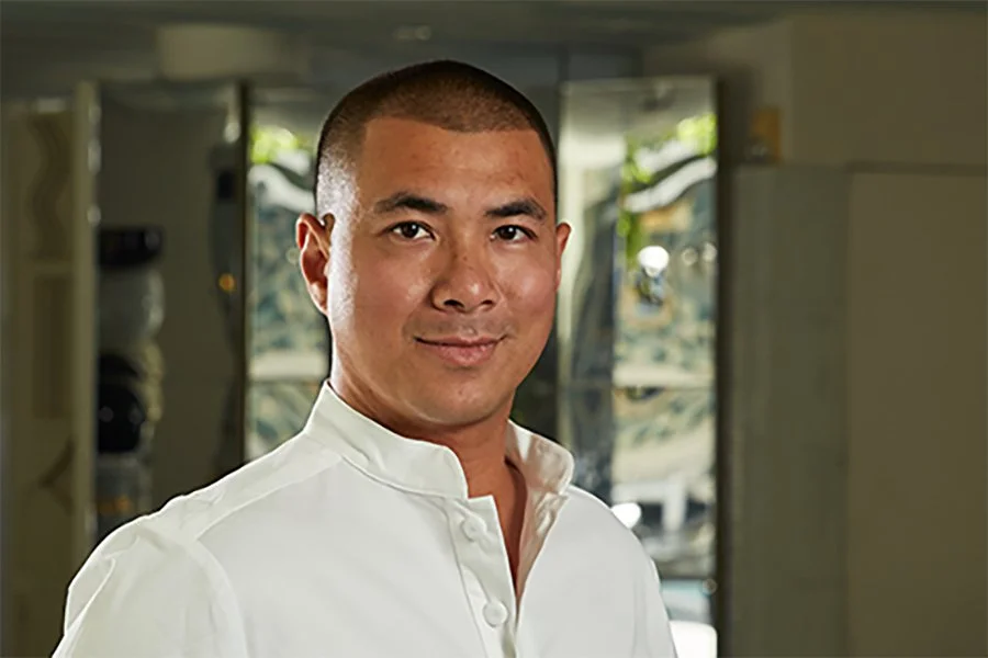 A man with a buzz cut wearing a white shirt, smiling indoors, with a boat visible in the background through an open door or window.