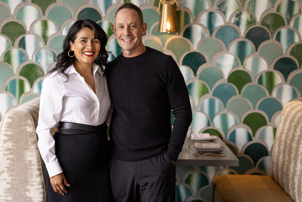 A smiling woman in a white blouse and black skirt and a smiling man in a black sweater standing together in a restaurant with colorful, patterned wallpaper in the background.