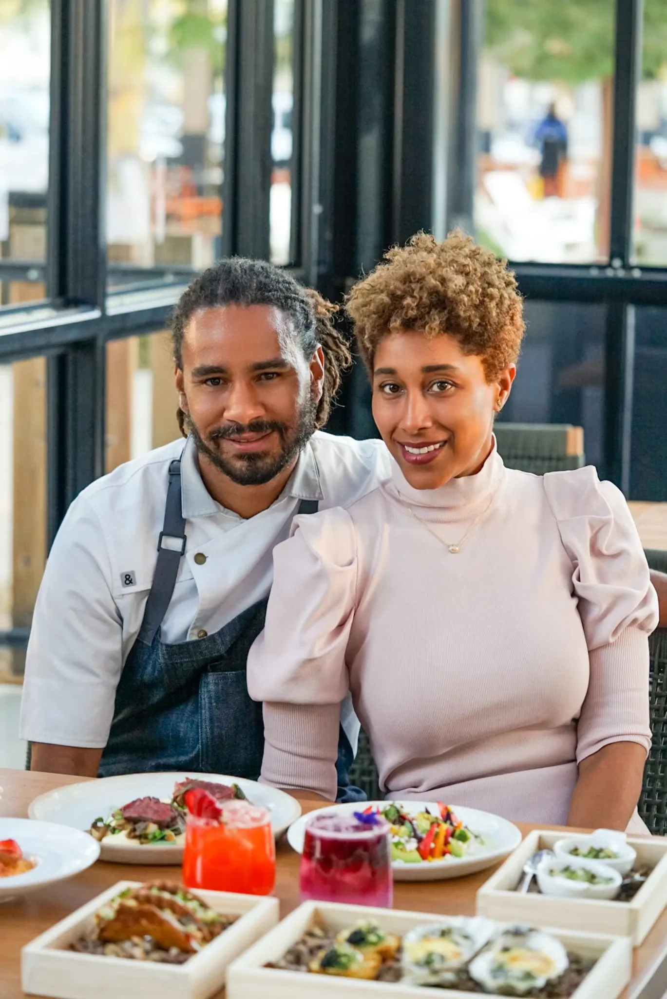 A woman and a man, both smiling, sitting at a table with various dishes and drinks in a restaurant.