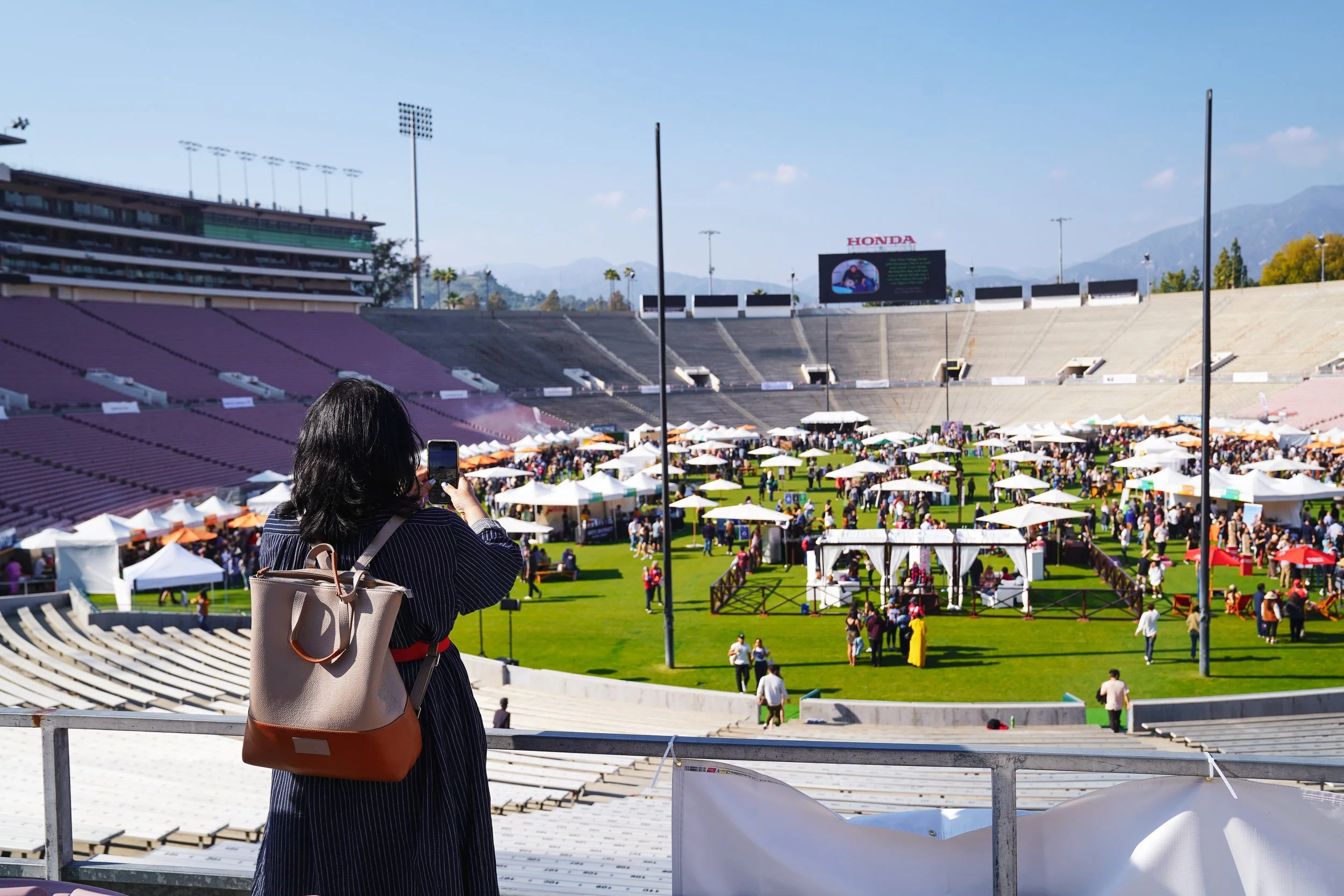 A woman taking a photo with her phone at the Rose Bowl stadium filled with white tents and people during an Masters of Taste on a sunny day.